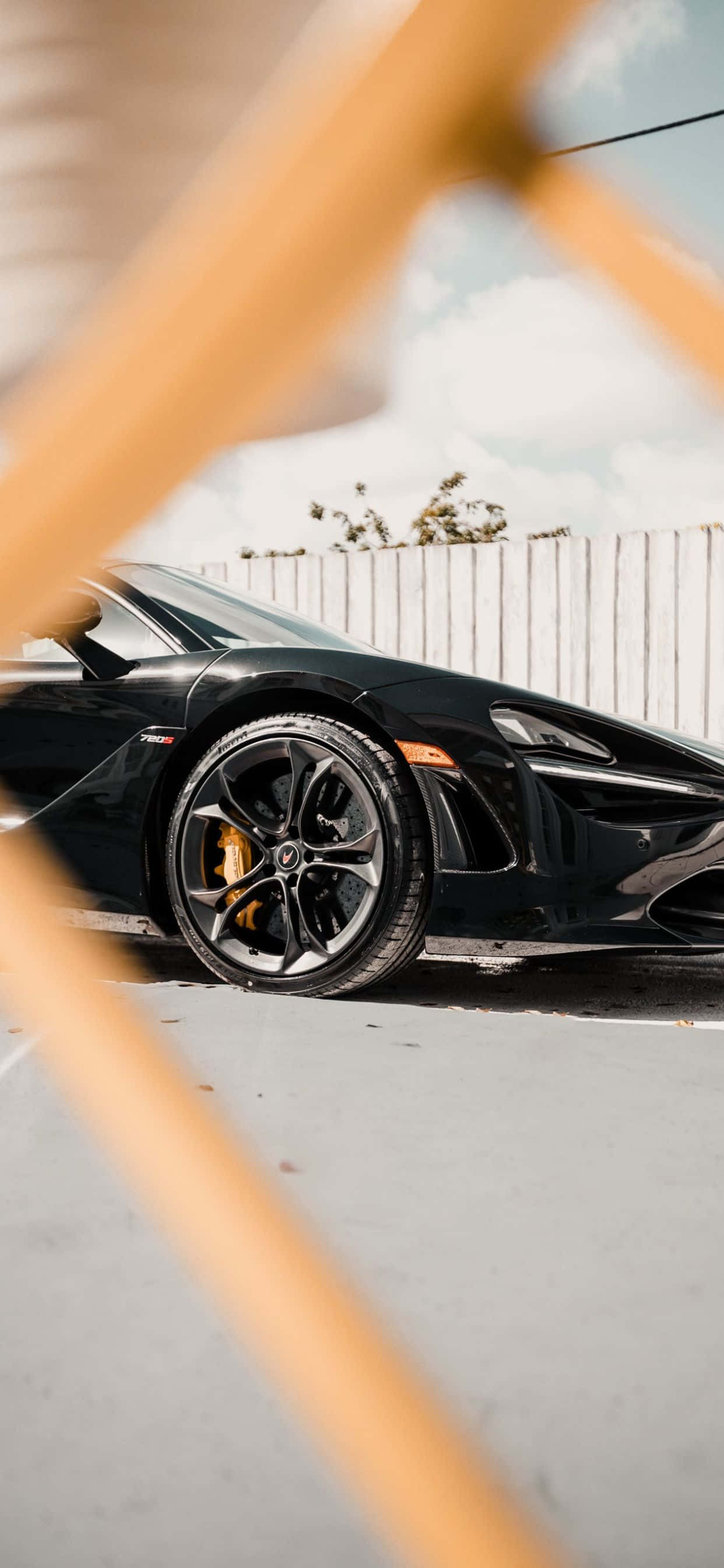 以 iPhone XS Max 與 McLaren 720S 開啟極致性能 壁紙 — 一輛時尚的黑色跑車，帶有黃色制動卡鉗，停在明亮的天空下，前景模糊的木樑框住了畫面。