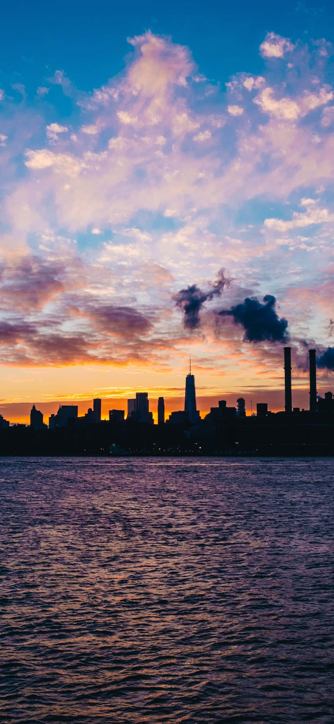 以 iPhone XS 感受紐約天際線的壯觀美景 壁紙 — 城市天際線在充滿活力的日落天空下呈現剪影，粉紅色、橙色和藍色的雲彩倒映在漣漪的水面上。