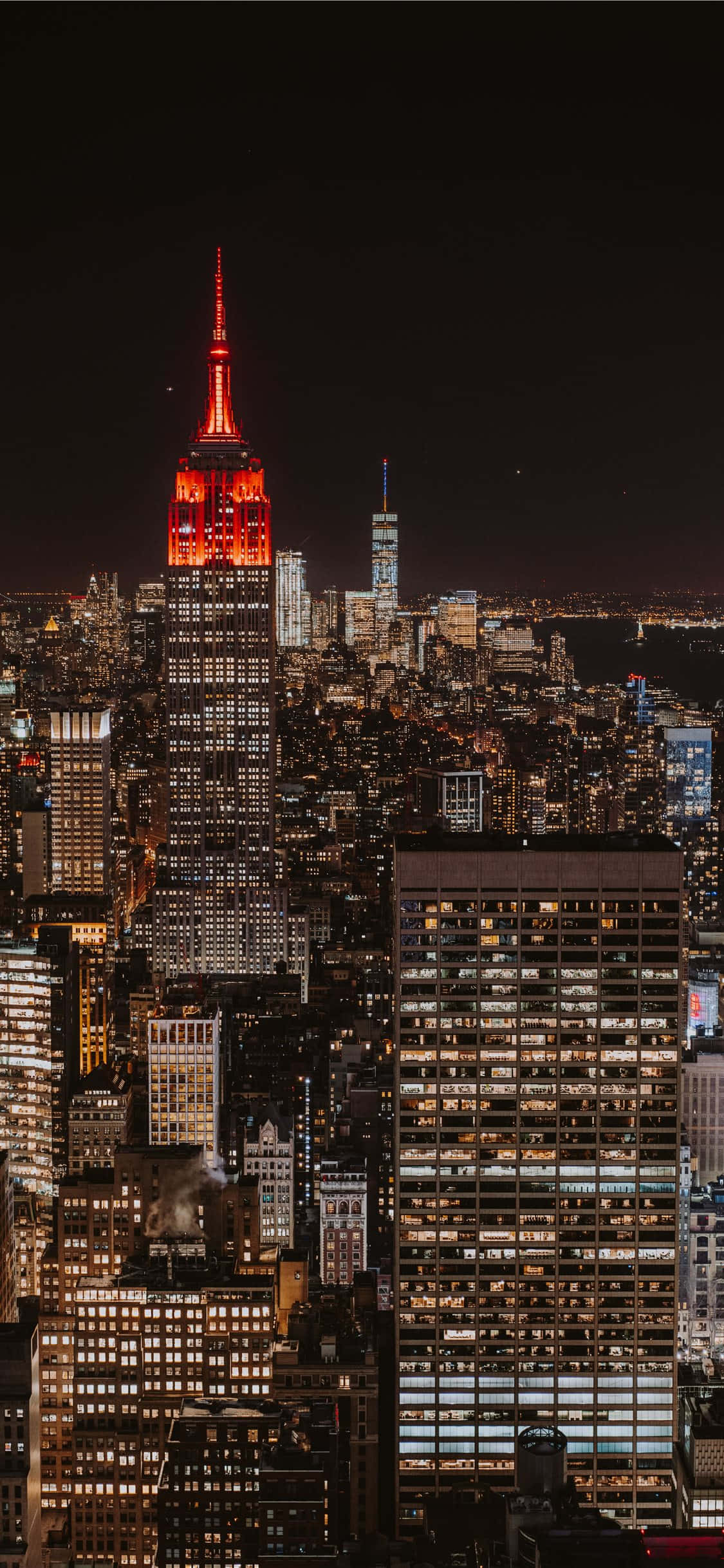 以 iPhone Xs 探索大蘋果 壁紙 — 紐約市天際線的壯麗夜景，以發光的紅色帝國大廈為主，周圍是無數燈火通明的摩天大樓。