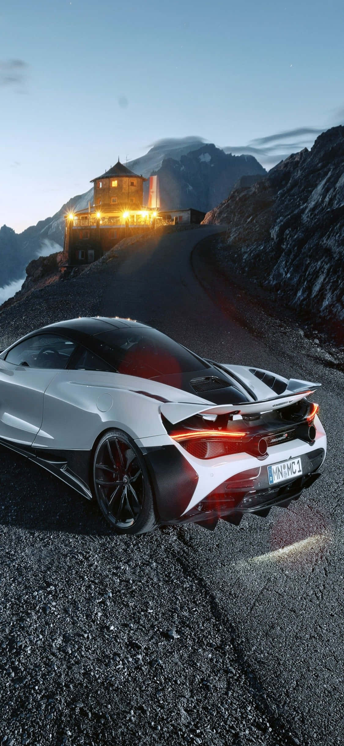 以 iPhone Xs 與 McLaren 720S 挑戰極限競速 壁紙 — 一輛流線型銀色跑車，帶有發光的紅色尾燈，停泊在黃昏時分蜿蜒的山路上，附近有一間燈火溫暖的阿爾卑斯小屋。