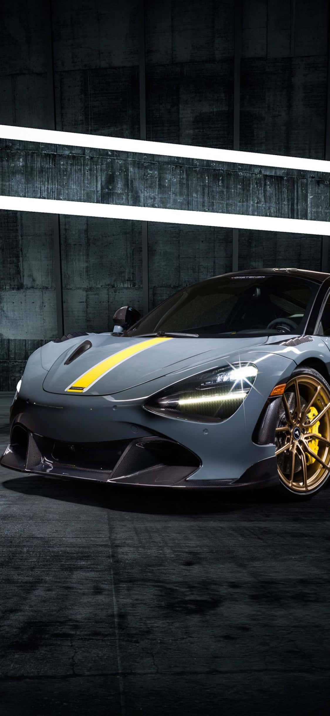 以 iPhone Xs 與 McLaren 720S 享受奢華旅程 壁紙 — 一輛時尚的灰色跑車帶有黃色點綴和金色輪圈，在明亮的白色燈光下展示，背景是深色混凝土牆。