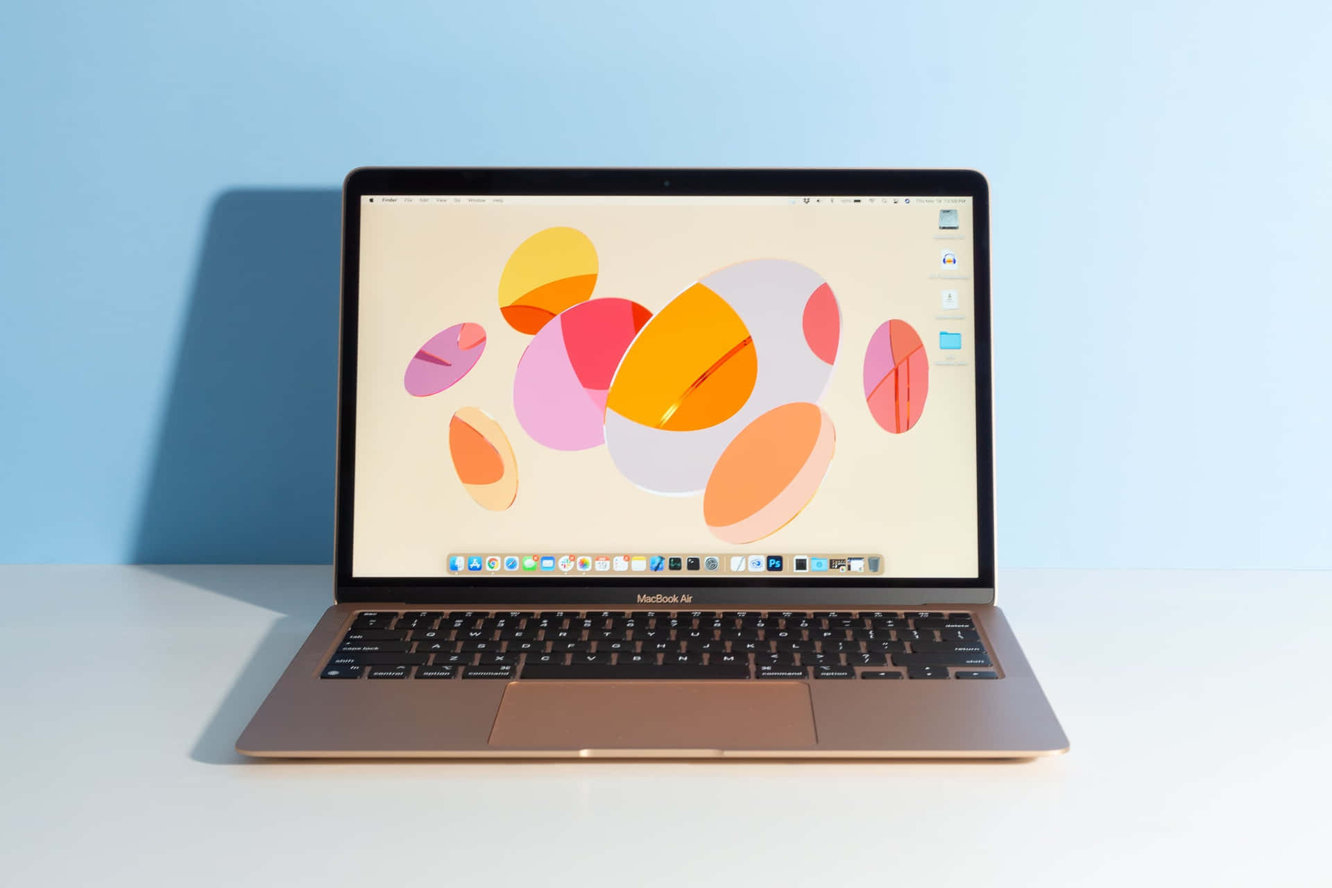 以 MacBook 將生產力提升至新境界 壁紙 — 一部纖薄的金色 MacBook Air 在白色表面上打開，對著柔和的藍色牆壁，顯示充滿活力的抽象桌布，上面有重疊的彩色圓圈。