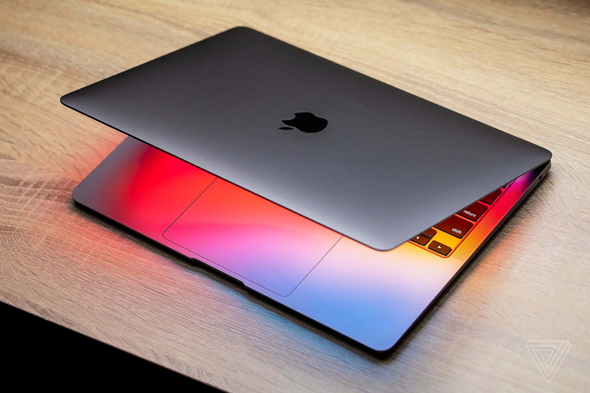 以 MacBook 獲得最頂級的便攜式計算體驗 壁紙 — 一個時尚的銀色MacBook蓋子微微打開，放在木桌上，露出鍵盤背光燈發出的鮮豔彩虹光芒。
