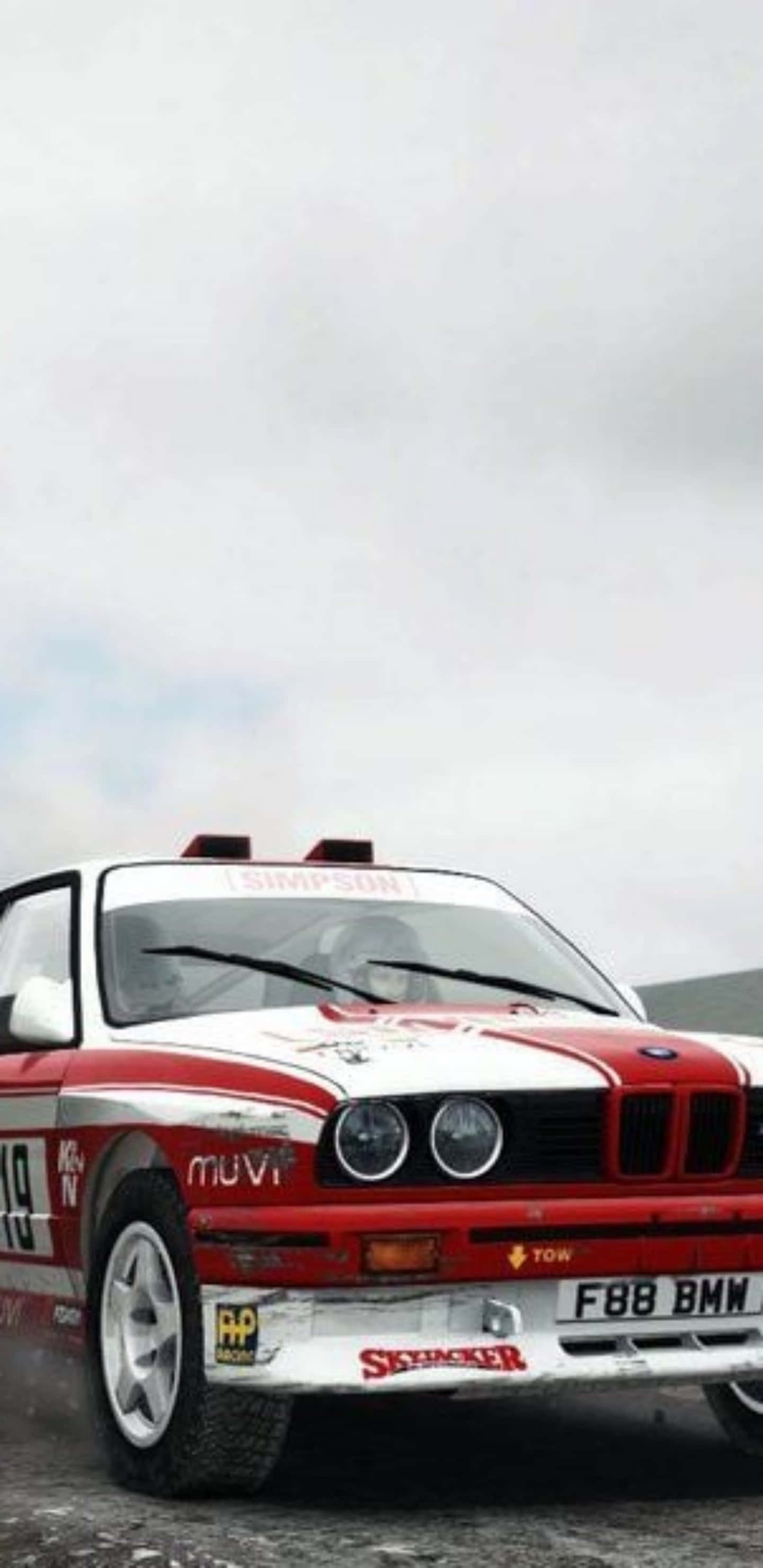 以 Pixel 3xl Dirt Rally 出擊，超越競爭對手 壁紙 — 一輛紅白相間的拉力賽車帶有贊助商貼紙，在多雲的天空下於濕滑道路上疾馳。