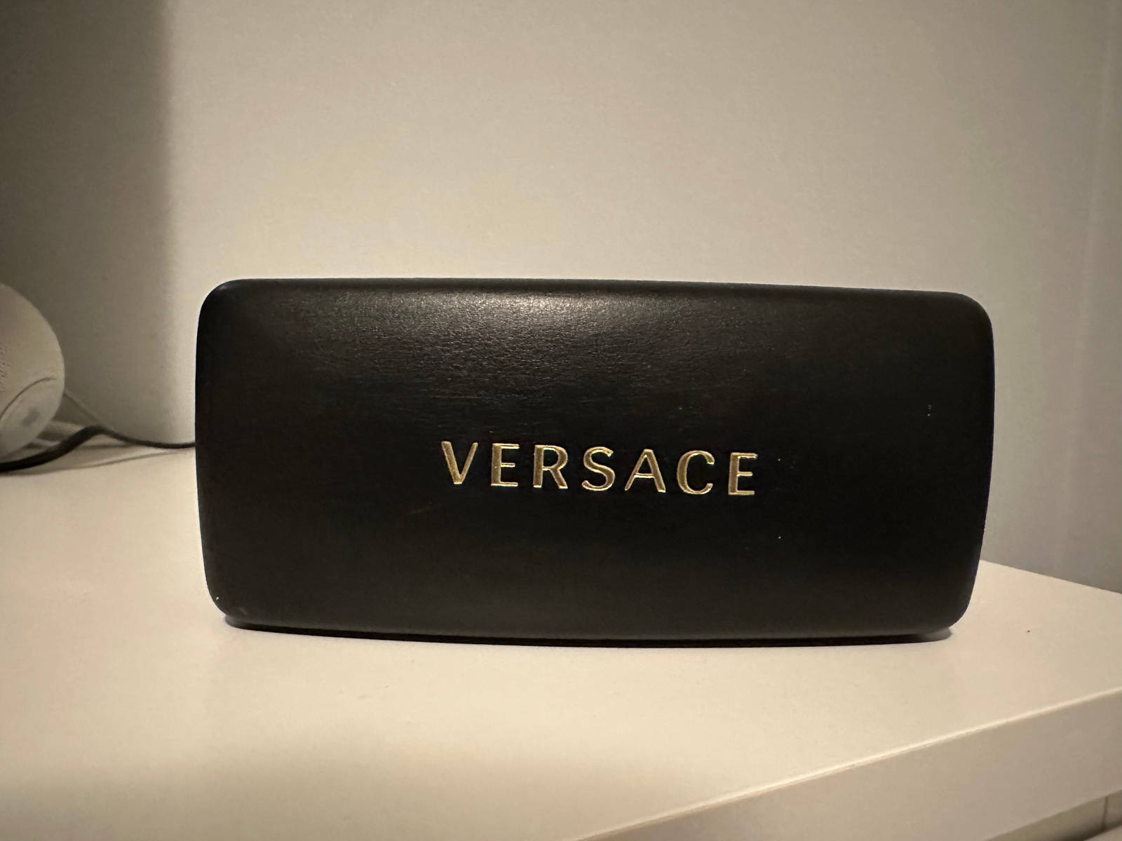 以 Versace 太陽眼鏡盒點亮你的造型 壁紙 — 一個時尚的黑色Versace眼鏡盒，帶有金色字樣，放在白色表面上，靠著素色牆壁。