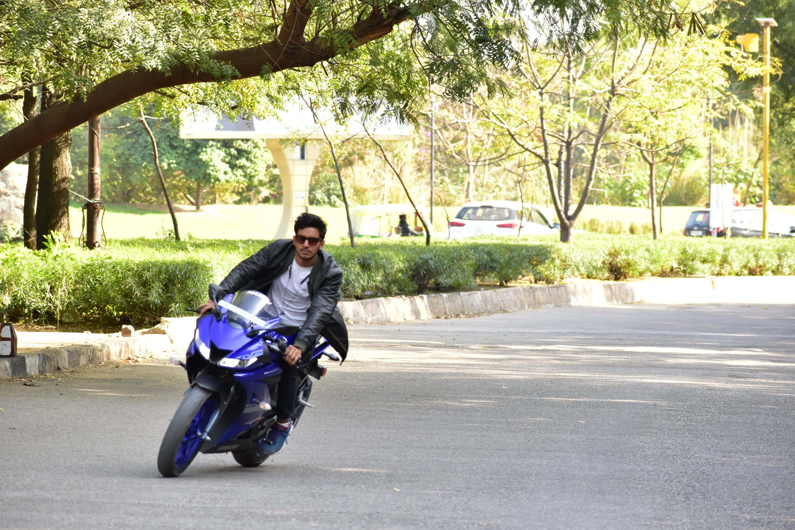 以 Yamaha R15 V3 將賽道推向高檔 壁紙 — 一名身穿黑色皮褸的男子，騎著一輛鮮藍色運動電單車，穿過陽光普照、綠樹成蔭、道路鋪設完善的公園。