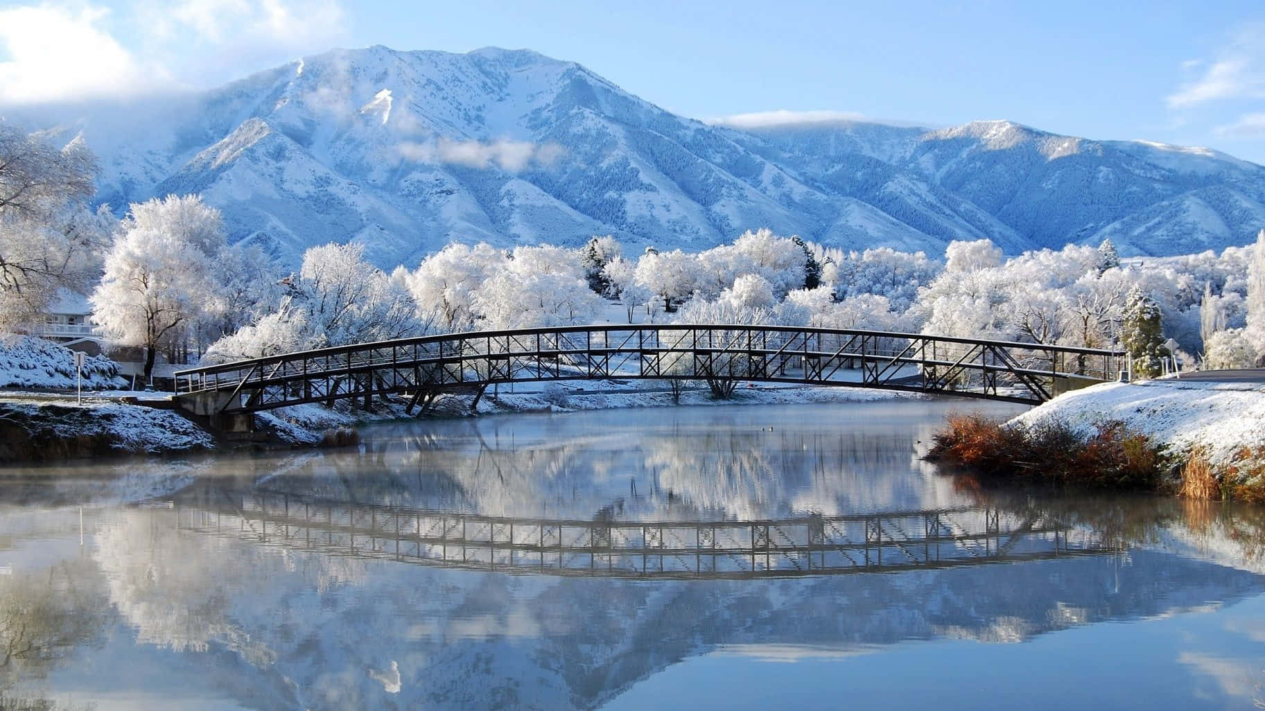 以1080p高畫質欣賞冬日風景 壁紙 — 一座被雪覆蓋的橋樑拱起在平靜的河面上，映照著白雪覆蓋的山脈和結霜的樹木，在清澈的藍天下。