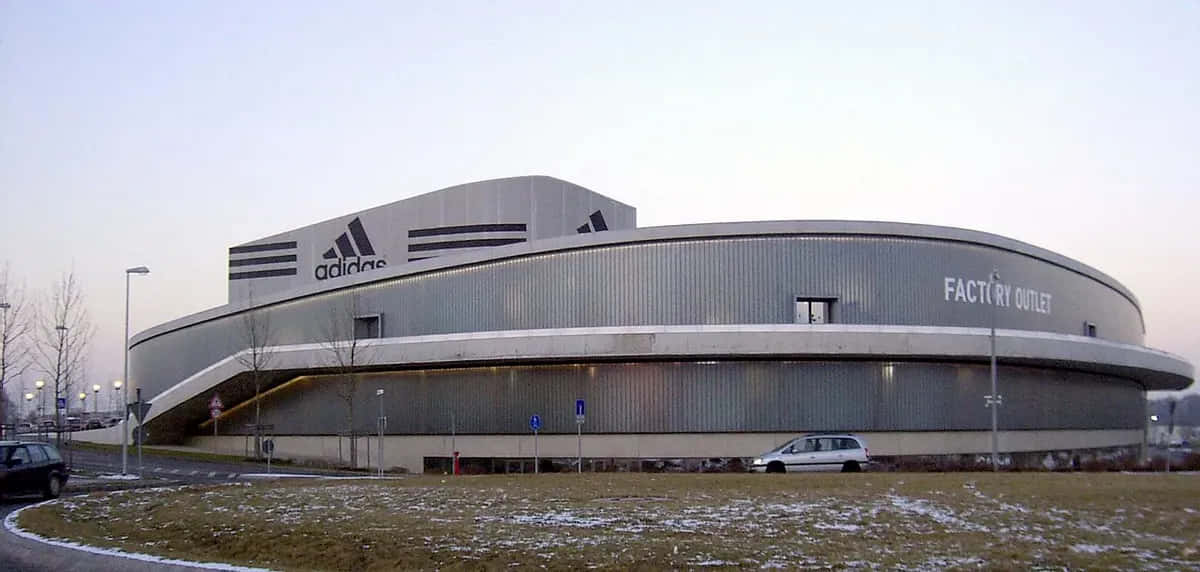 以Adidas刻下你的時尚印記 壁紙 — 一座大型弧形 adidas 工廠直銷建築，帶有金屬壁板和前面有雪塵的草地，在黃昏時分。