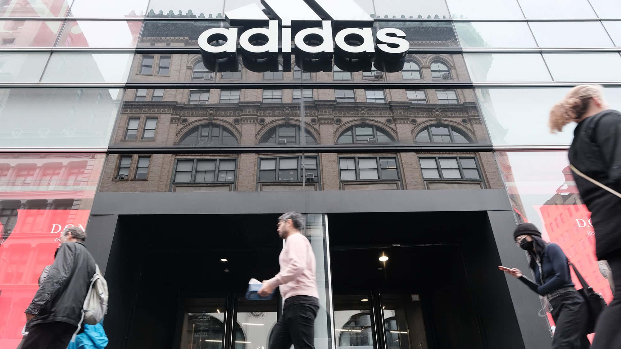 以Adidas運動服裝提升你的表現 壁紙 — 行人走過一間現代化的adidas商店，玻璃外牆反射出背後歷史悠久的磚建築。