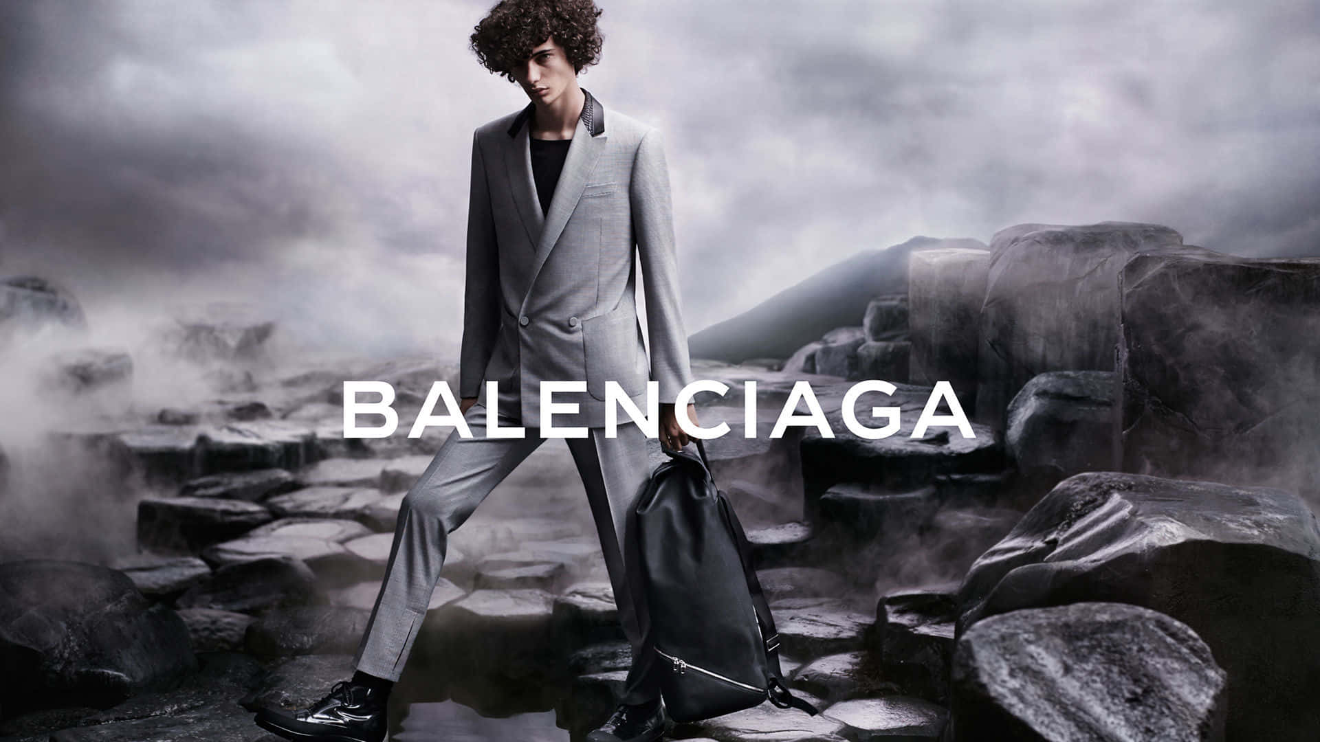 以Balenciaga提升您的街頭時尚品味 壁紙 — 一位身穿灰色西裝的模特兒在風暴天空下自信地走過霧氣瀰漫的岩石地形，並帶有 'BALENCIAGA' 文字疊加。