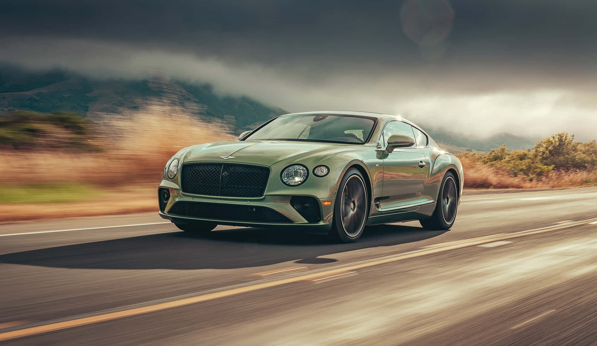 以Bentley Sport衝向巔峰 壁紙 — 一輛時尚的綠色賓利 Continental GT 在戲劇性的多雲天空下沿著蜿蜒的山路飛馳。