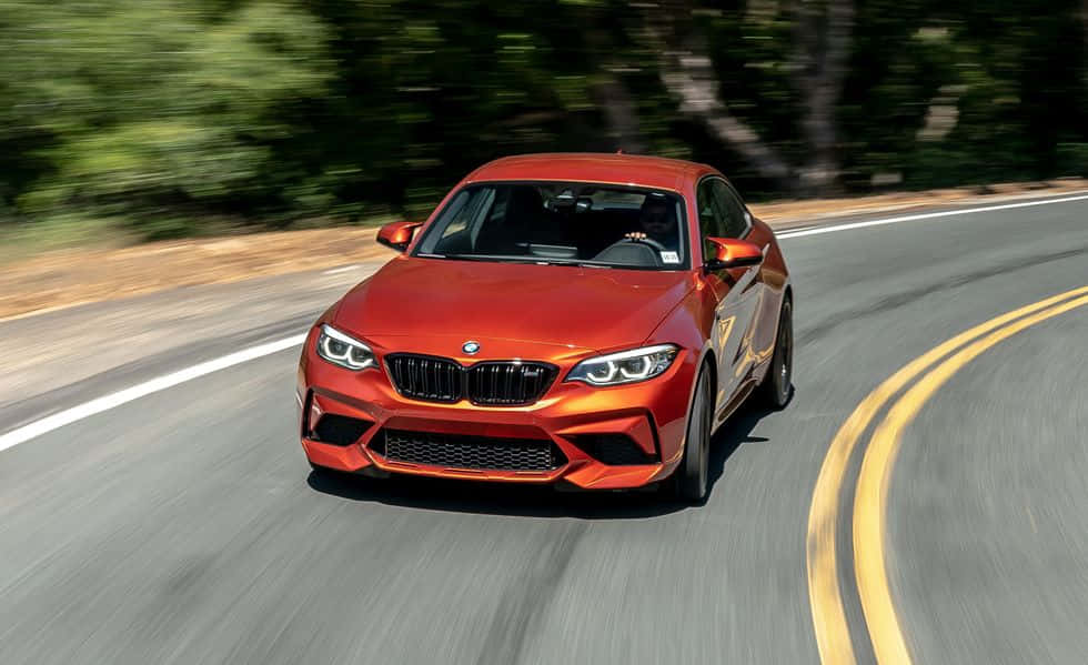 橙色BMW M2 Coupe在彎曲的道路上行駛，有綠色樹木和黃色線條。