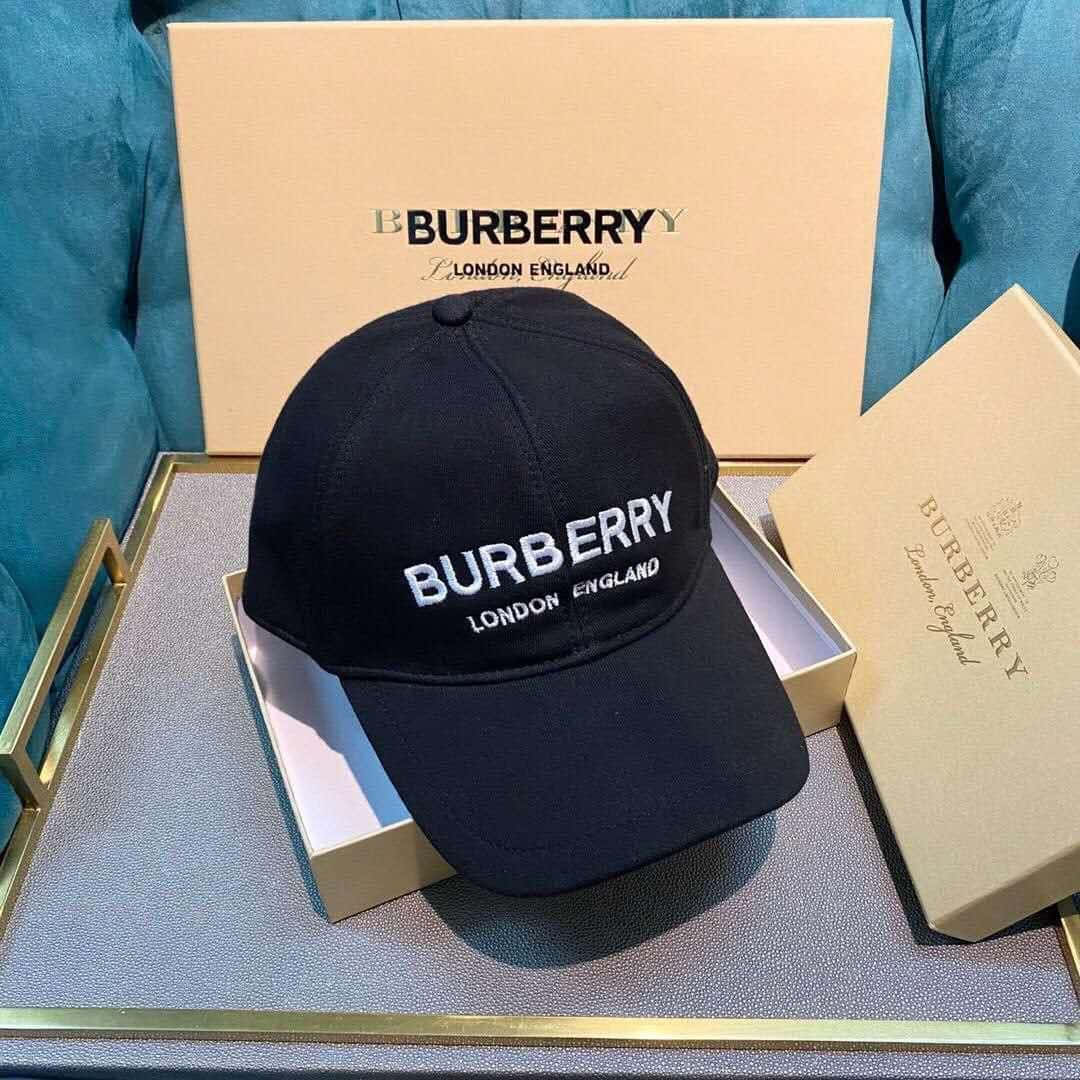 以Burberry展現高雅風格 壁紙 — 一頂帶有白色標誌的黑色Burberry帽子優雅地放在一個米色盒子裡，置於金色邊緣的托盤上，背景是藍綠色天鵝絨。