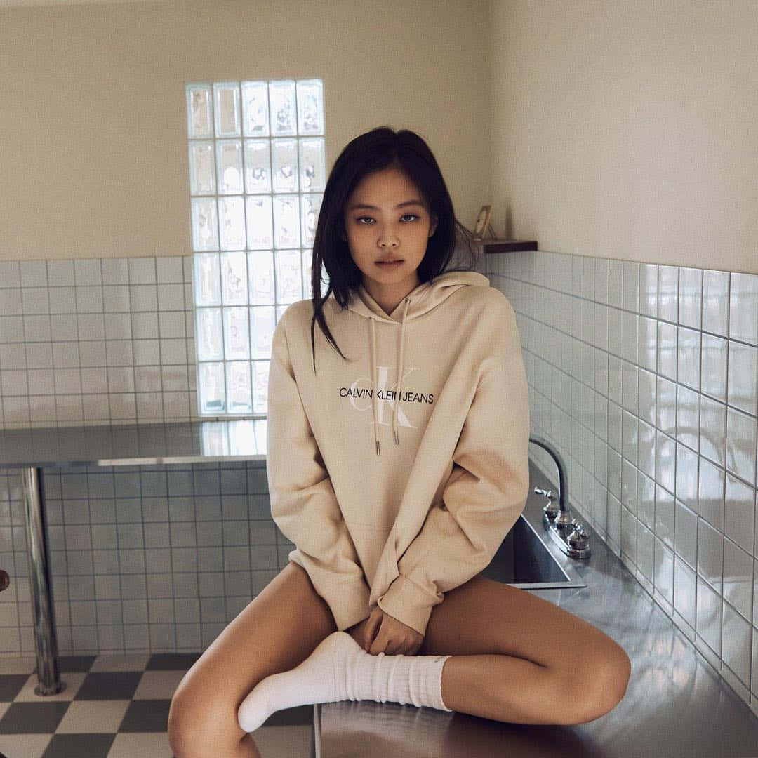 以Calvin Klein保持時尚品味 壁紙 — 一位身穿米色連帽衫的年輕女子，平靜地坐在瓷磚浴室的鋼製水槽上，浴室有棋盤地板和玻璃磚窗。