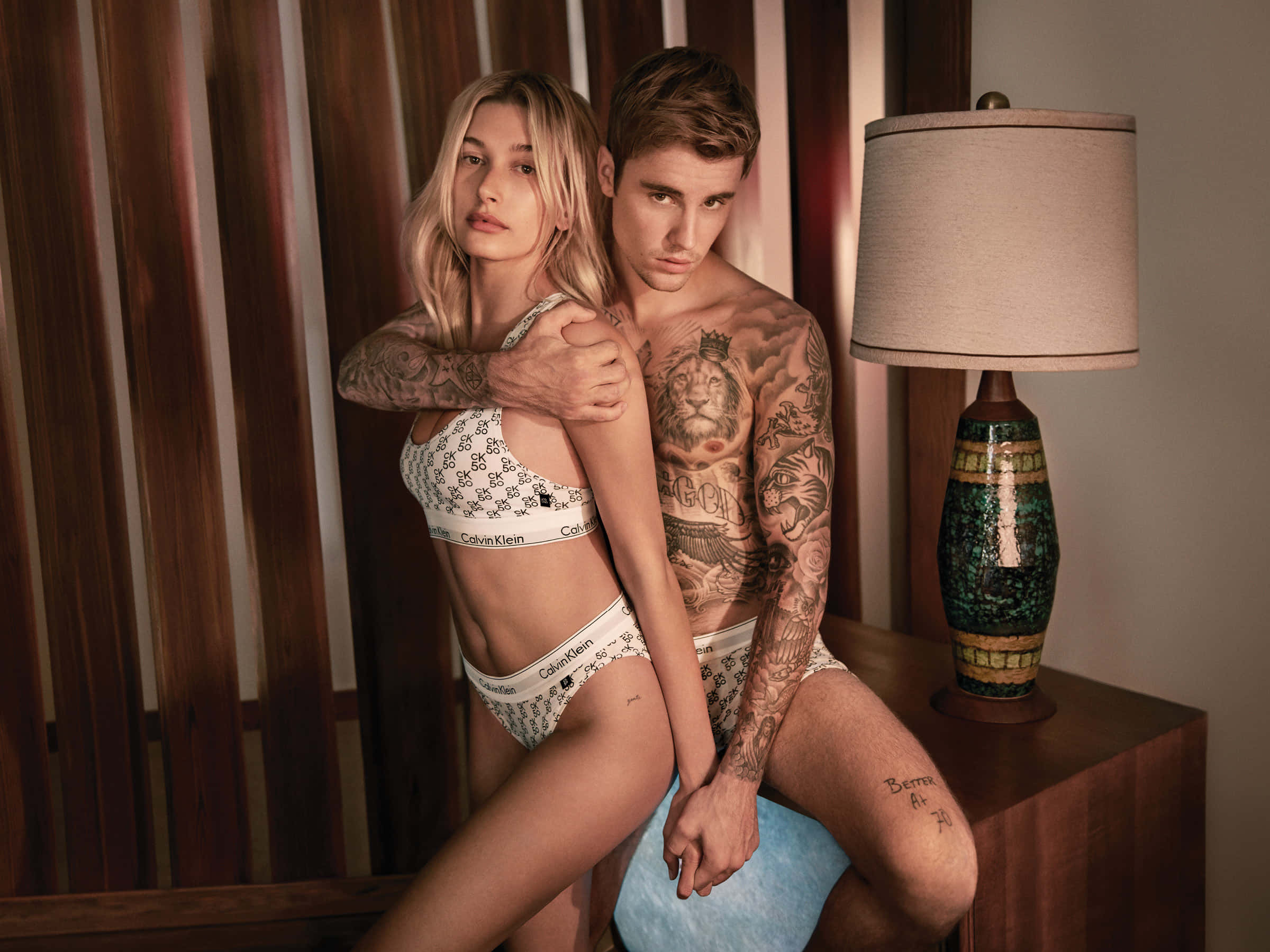 以Calvin Klein展現個人風格 壁紙 — 一對紋身情侶穿著Calvin Klein內衣，在溫暖照明的木質房間裡，與圖案燈具親密擺姿勢。