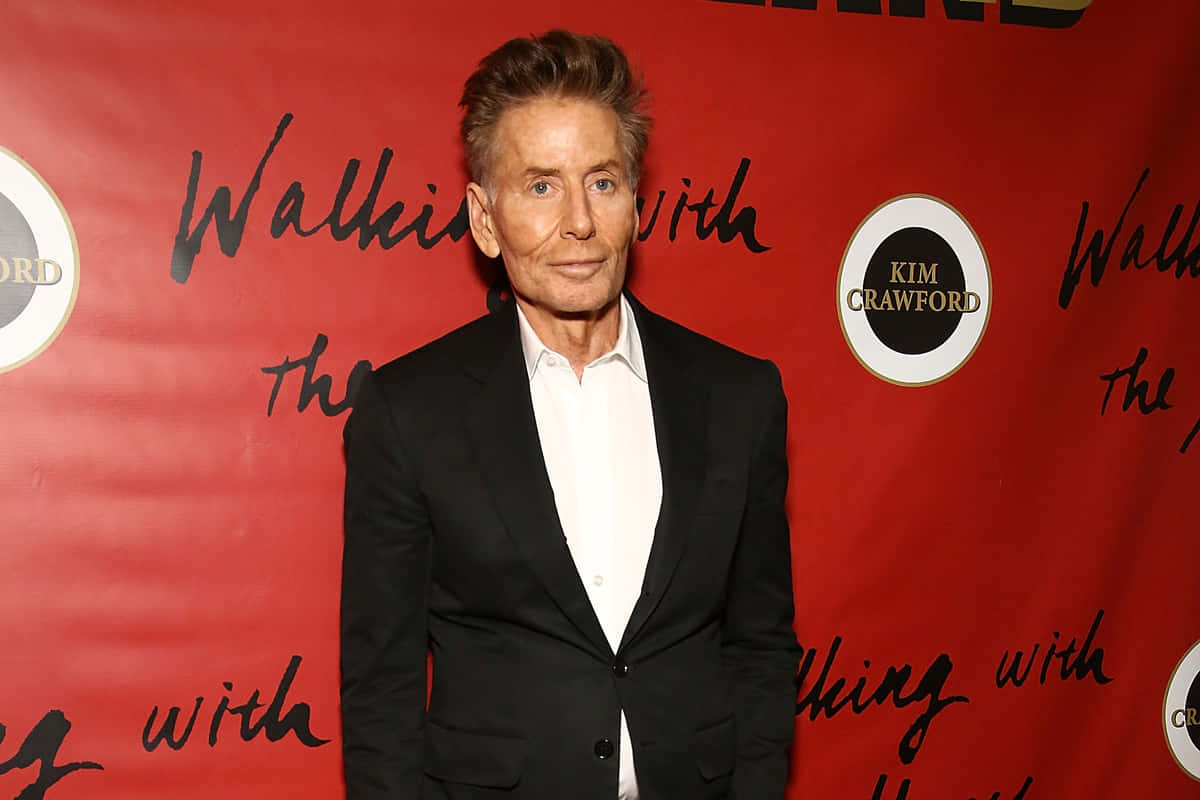 以Calvin Klein展現大膽自我 壁紙 — 一名身穿黑色西裝和白色襯衫的男子自信地站在紅色背景前，背景上寫有 'Walking with Kim Crawford' 字樣。