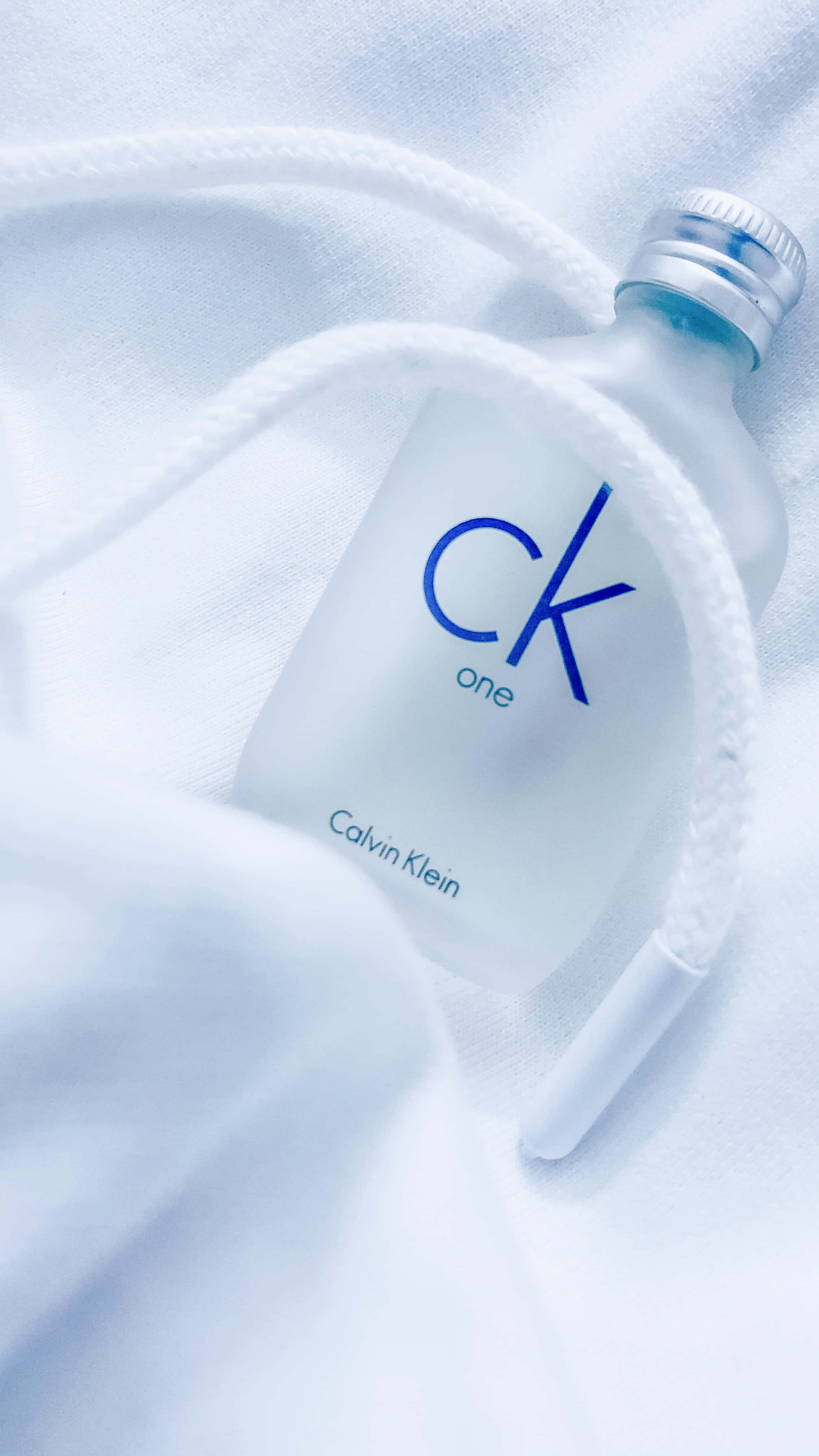 以Calvin Klein感受自信與性感 壁紙 — 一個磨砂玻璃 Calvin Klein CK One 香水瓶放在柔軟的白色布料上，白色繩索纏繞著。