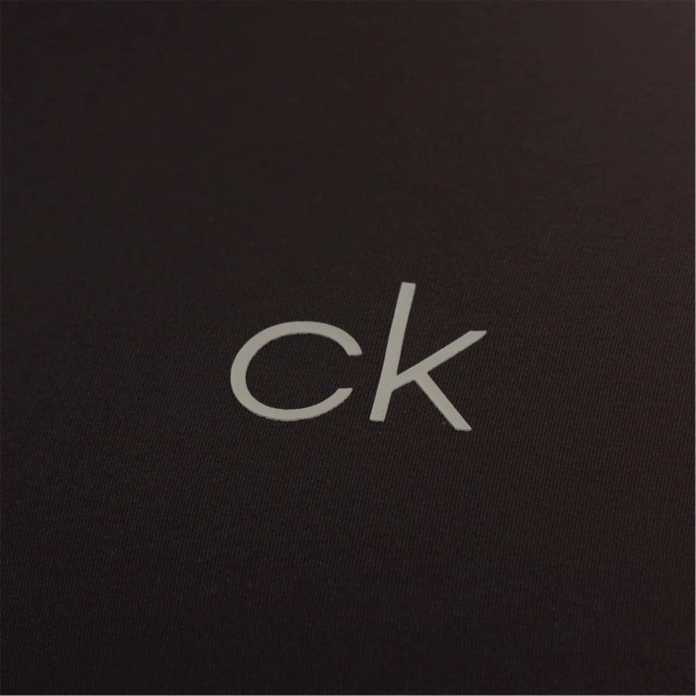 以Calvin Klein迎接延長季節 壁紙 — 極簡風格特寫，銀色 'ck' 標誌位於有紋理的黑色布料上，光線柔和。