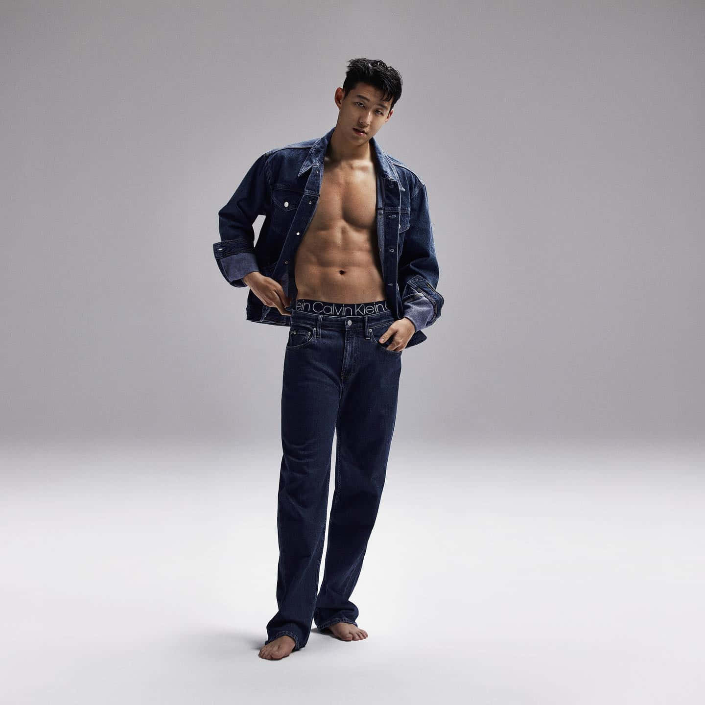 以Calvin Klein重新定義自我 壁紙 — 一個肌肉發達的男人穿著開著的牛仔外套和牛仔褲，赤腳站在純色攝影棚背景前。
