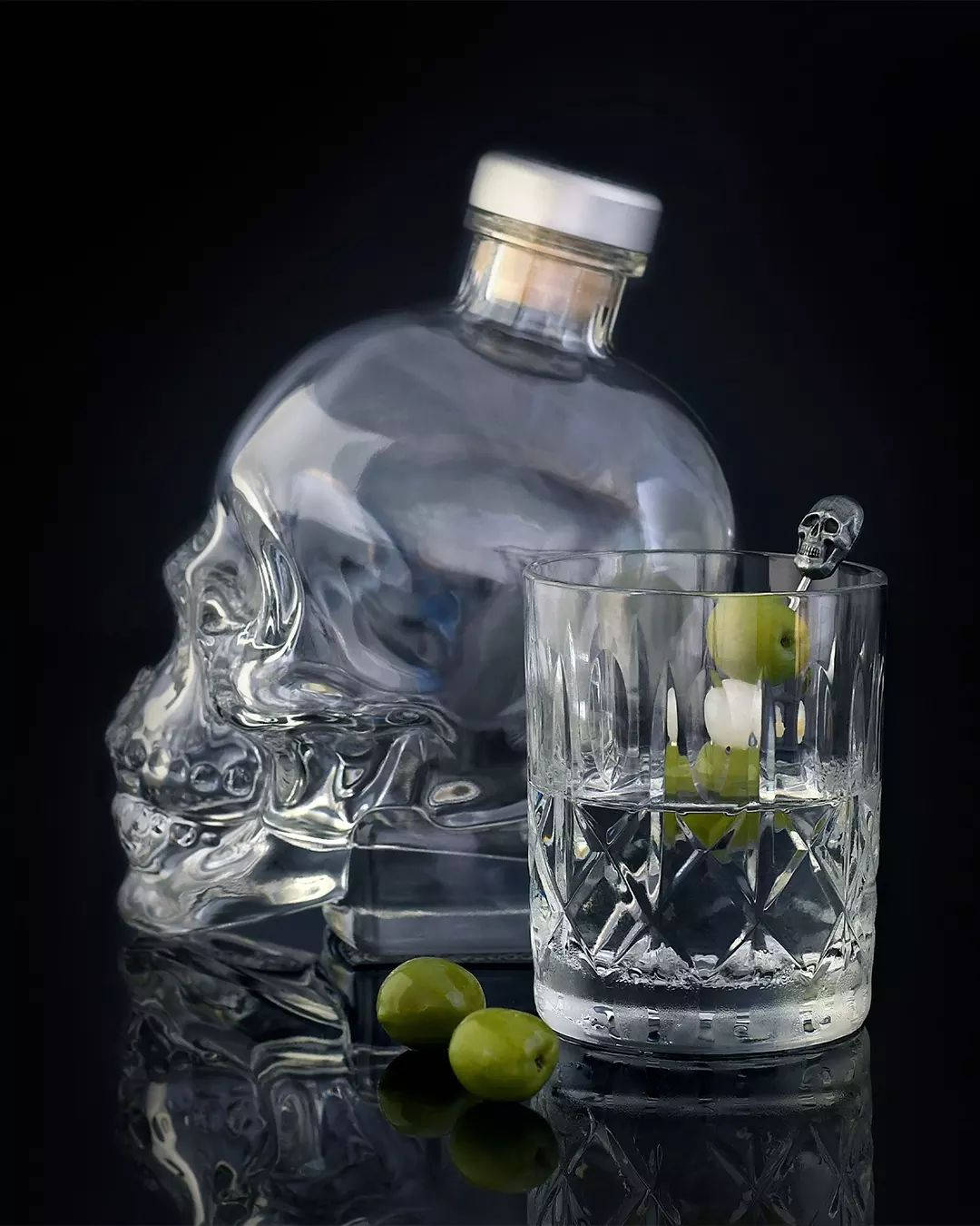 骷髏伏特加酒瓶和帶橄欖的玻璃杯在黑色反光表面上。