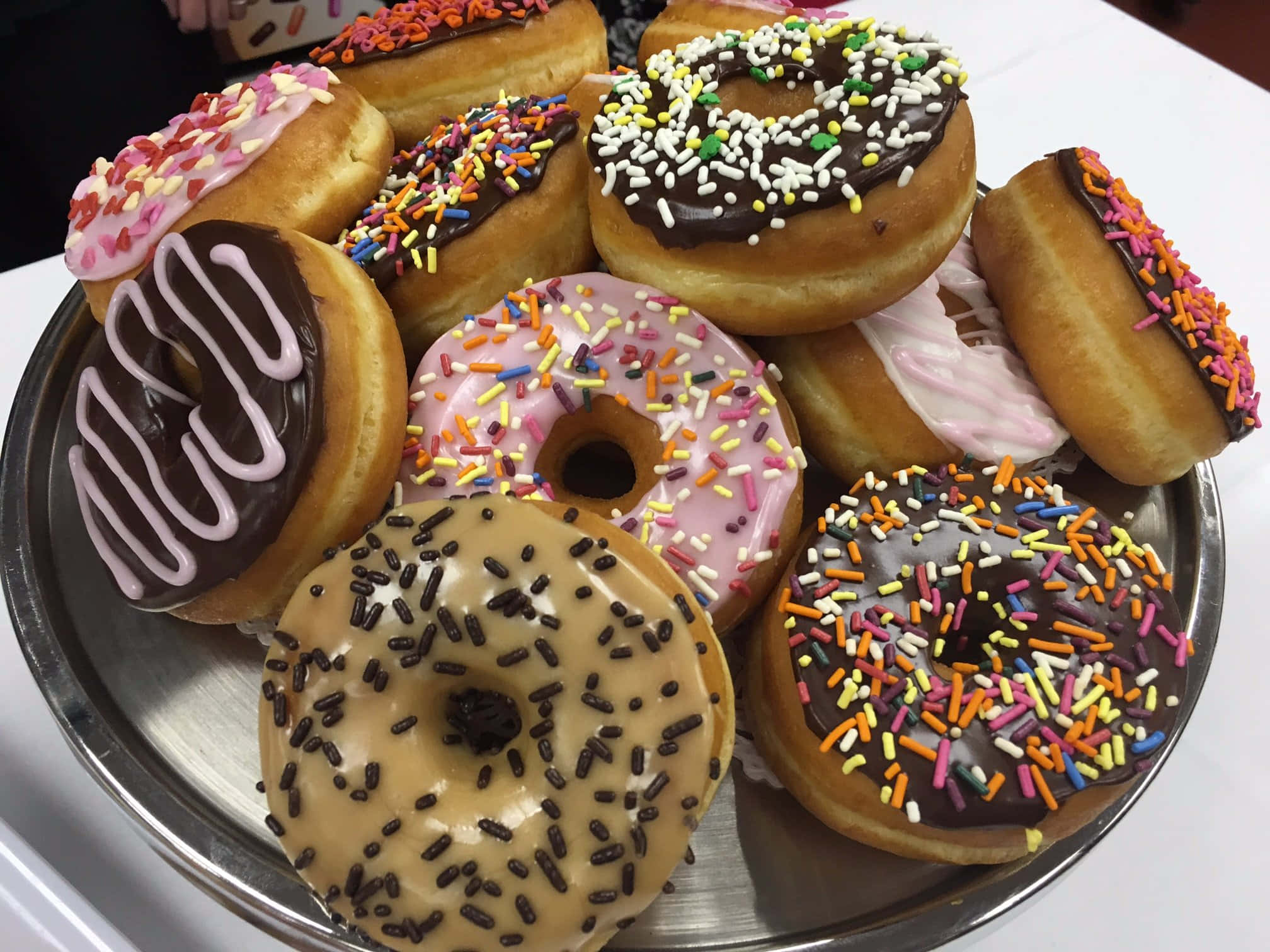 以Dunkin' Donuts補充每日咖啡因 壁紙 — 一個金屬托盤上堆滿了各式各樣的釉面甜甜圈，上面撒滿了彩色糖粒和朱古力醬。