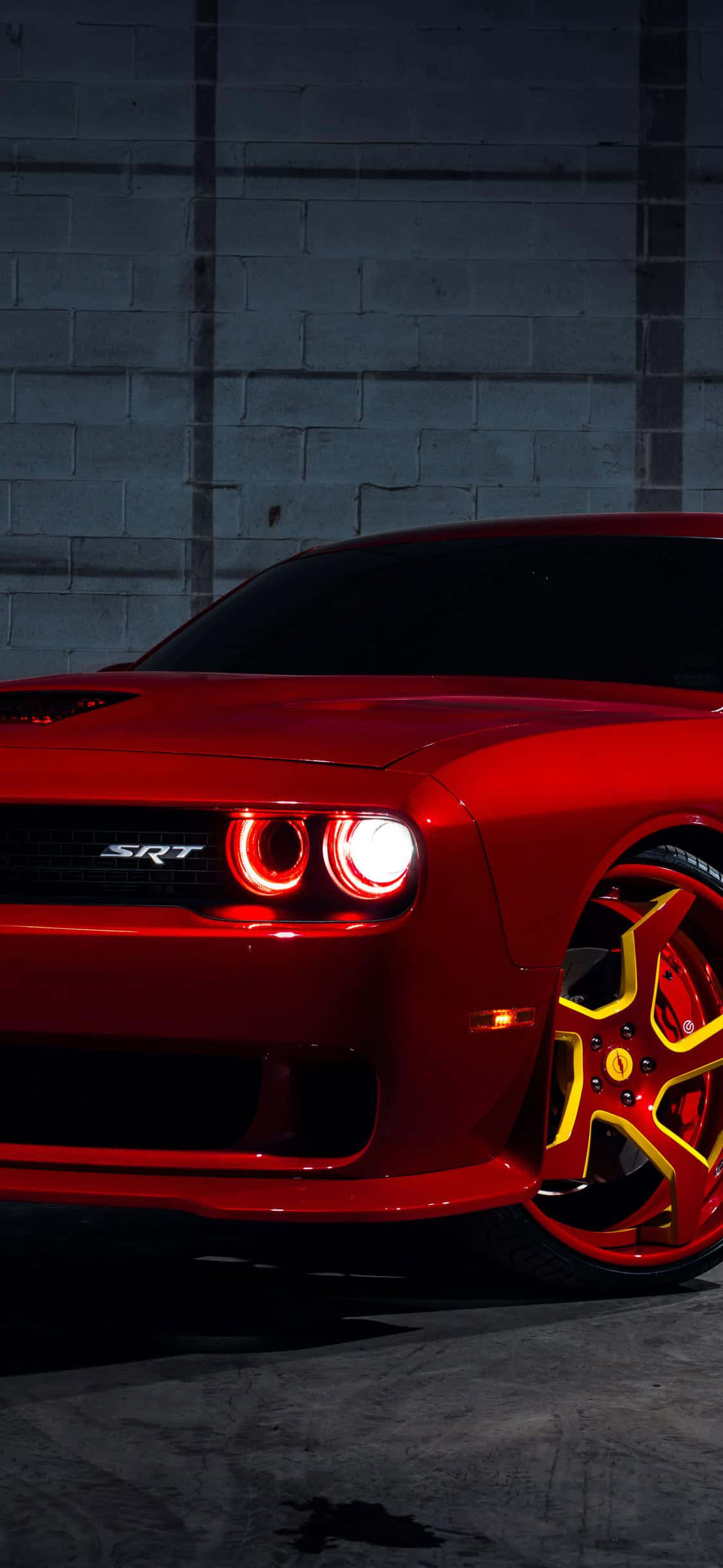 以Hellcat iPhone提升移動體驗 壁紙 — 一輛光澤的紅色Dodge Challenger SRT，帶有發光的頭燈和黃色點綴的輪圈，停在深色混凝土牆前。