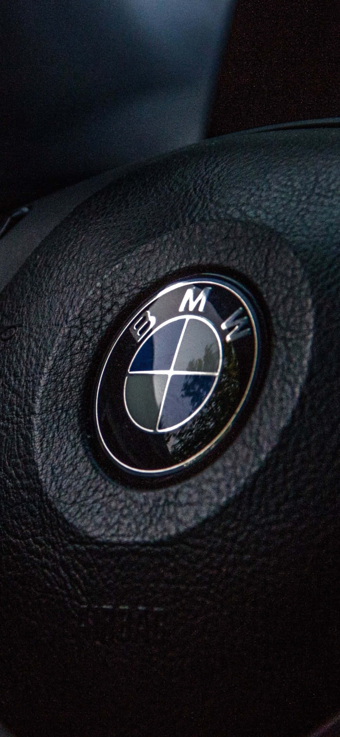 以iPhone X與BMW體驗極致奢華 壁紙 — 黑色皮革軚環上帶有反射性鍍鉻飾邊的BMW標誌特寫。
