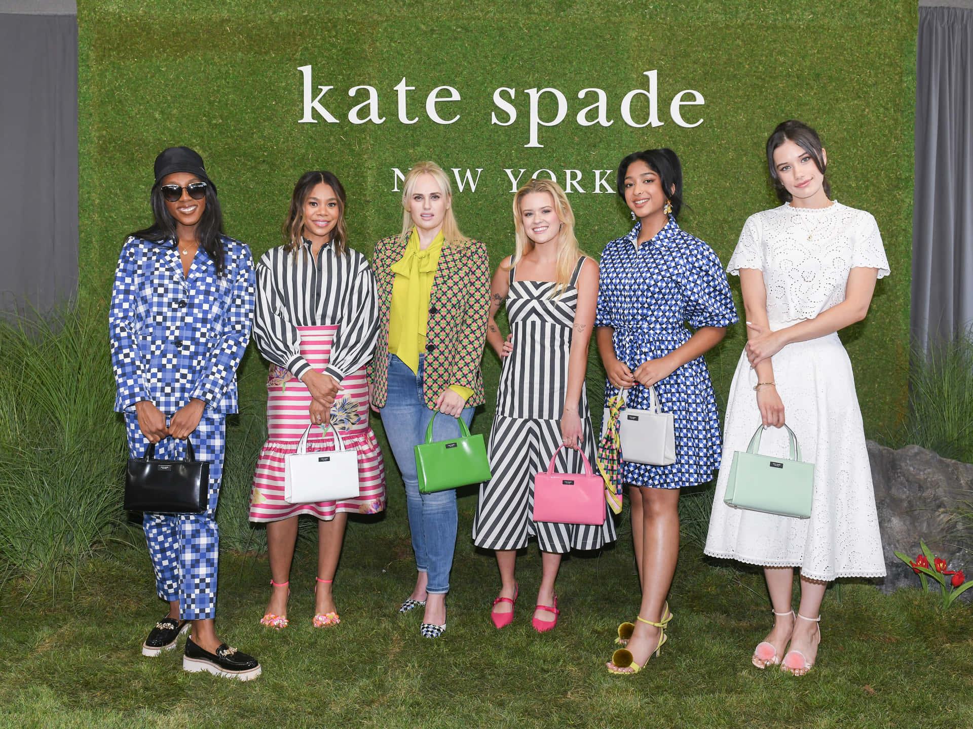 六位穿著時尚服裝和手袋的女性在草地上參加 Kate Spade 活動時擺姿勢。