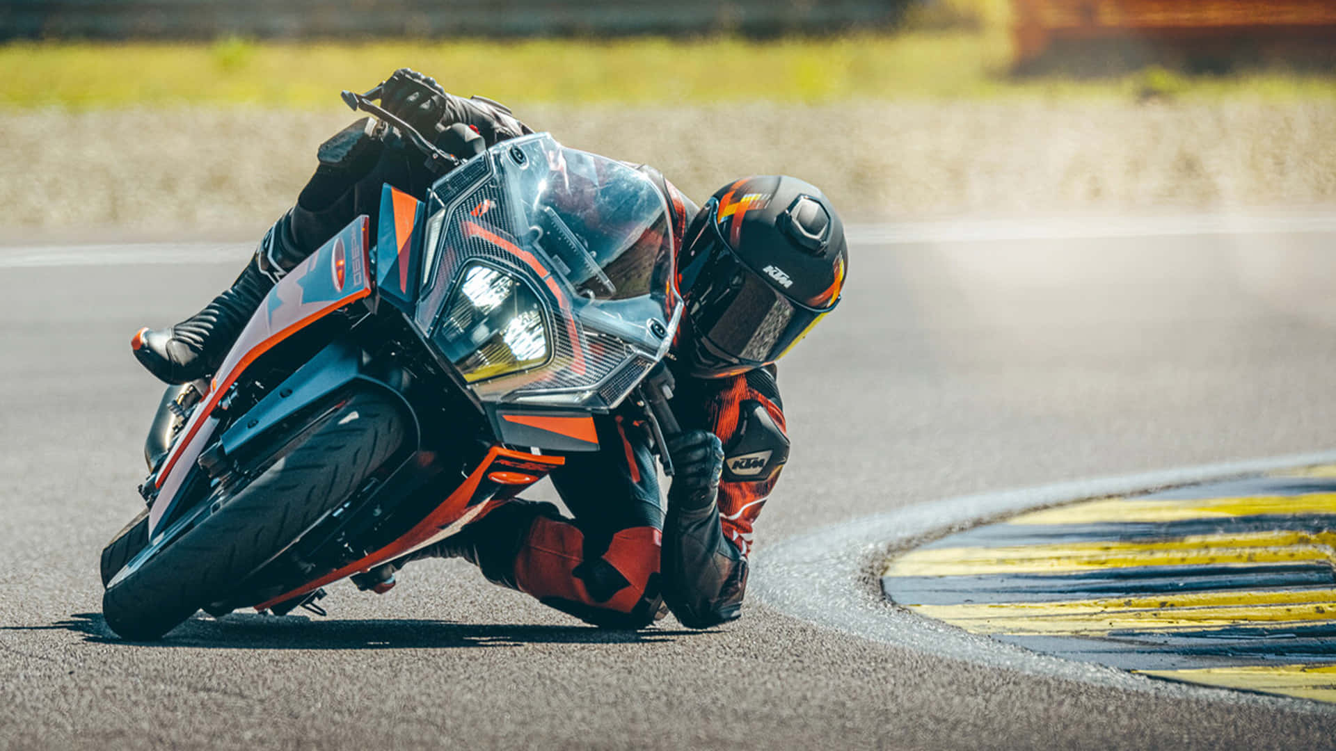 以KTM Duke征服街道 壁紙 — 一名電單車手在陽光明媚的賽道上，穿著鮮豔的橙色和黑色裝備，急劇傾斜入彎。