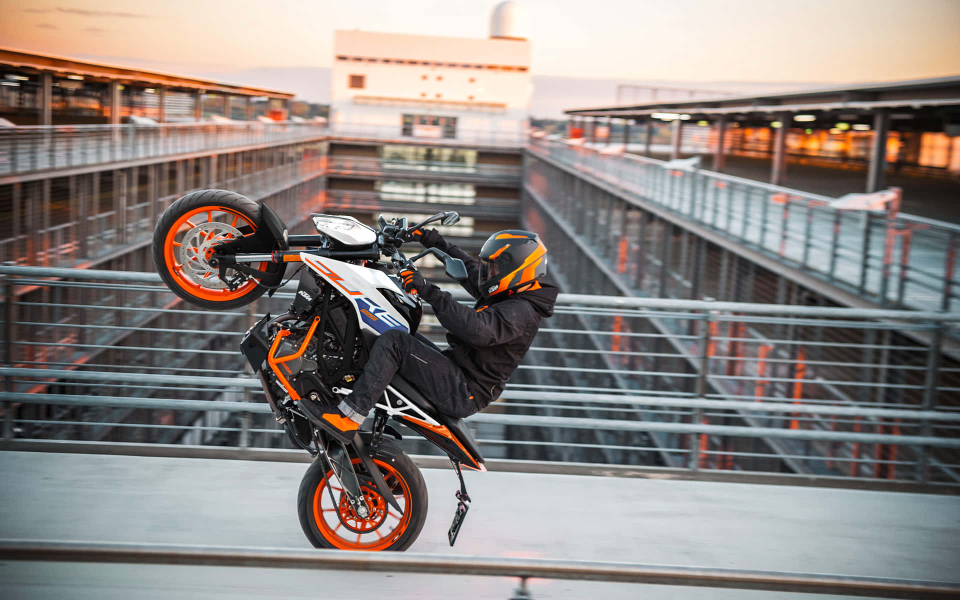以KTM Duke提升城市駕馭體驗 壁紙 — 一名電單車手在日落時分，於金屬橋上騎著橙色和白色的電單車表演前輪離地。