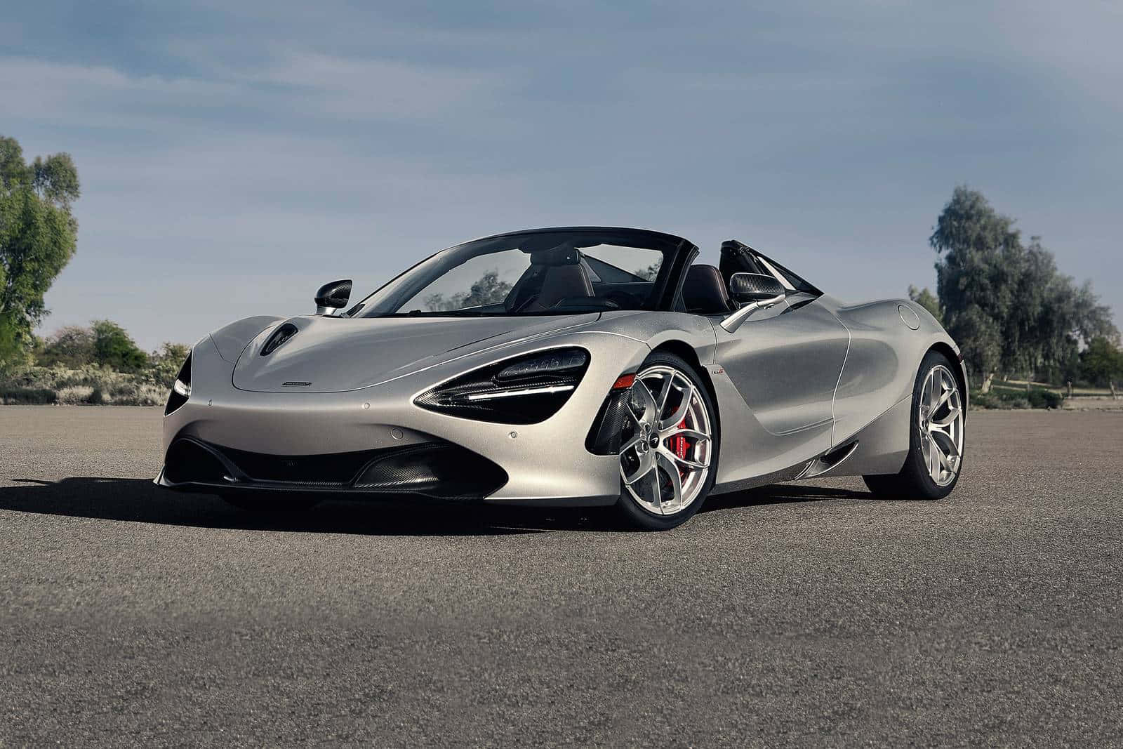 銀色McLaren 720S Spider開篷跑車停在鋪砌的道路上，有樹木和天空。
