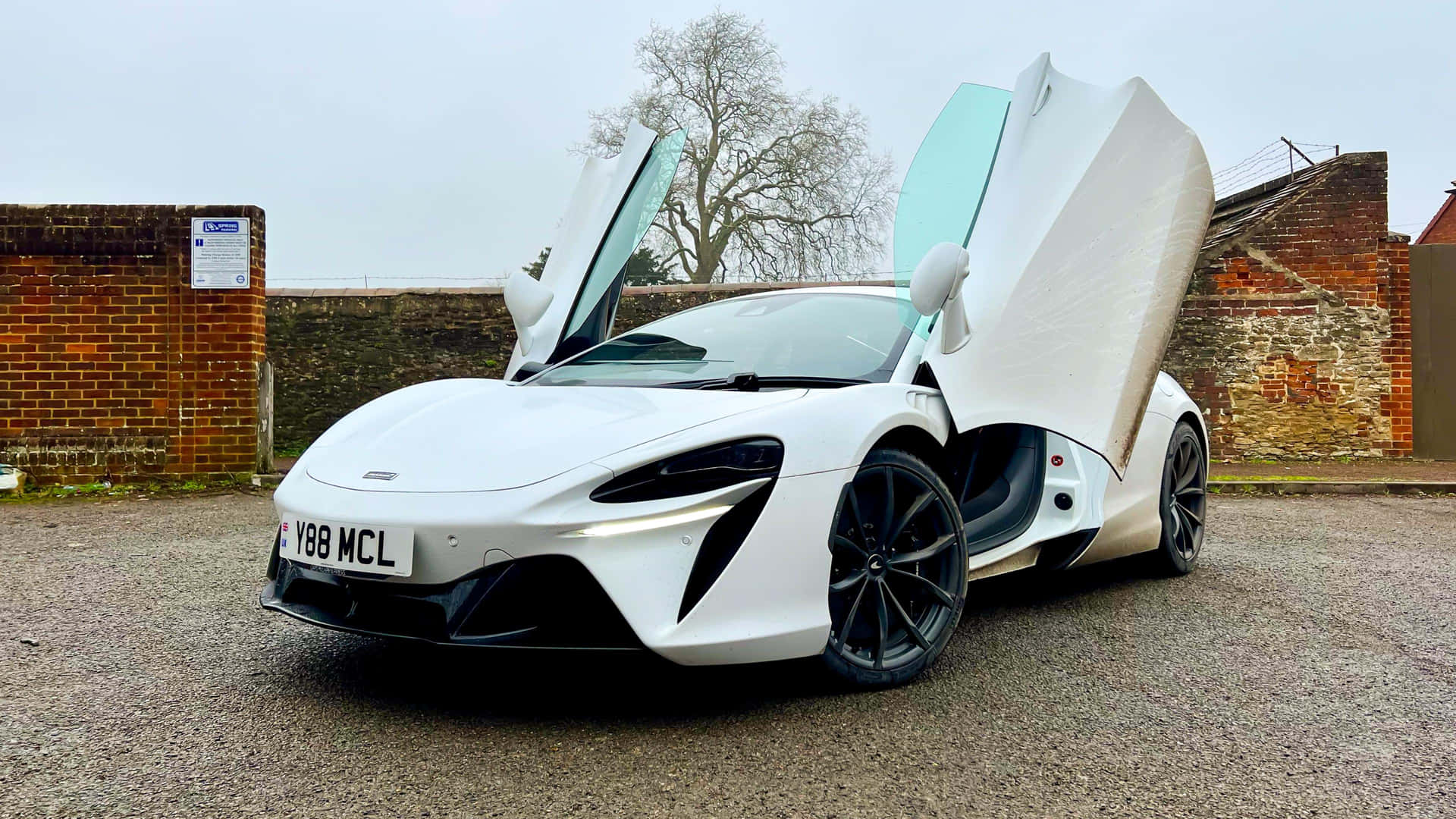 白色McLaren跑車，鷗翼門打開，停在磚牆附近的碎石地上。