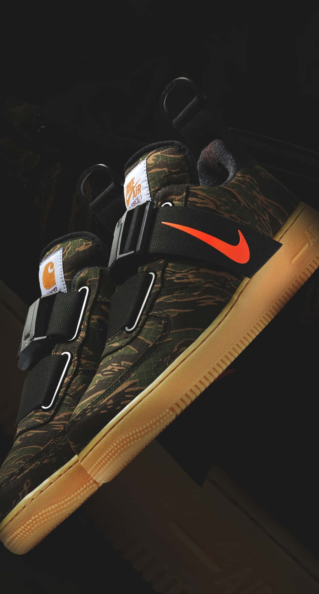 迷彩 Nike Carhartt 運動鞋，帶有橙色 swoosh 標誌和棕褐色鞋底，置於深色背景前。
