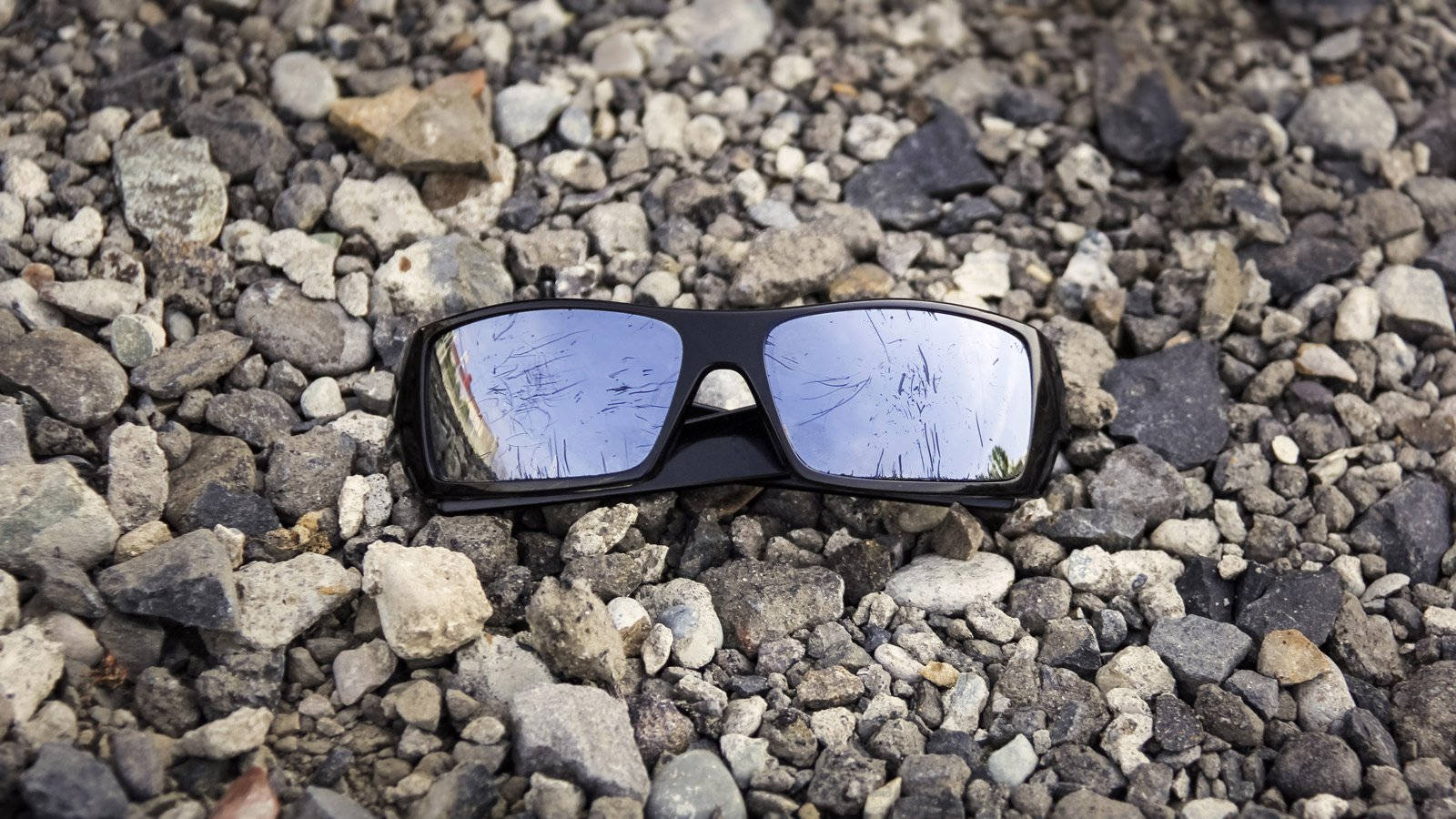 以Oakley運動健行太陽眼鏡展現冒險精神 壁紙 — 一副黑色鏡框、鏡面有刮痕的太陽眼鏡擱在灰色和白色碎石上。