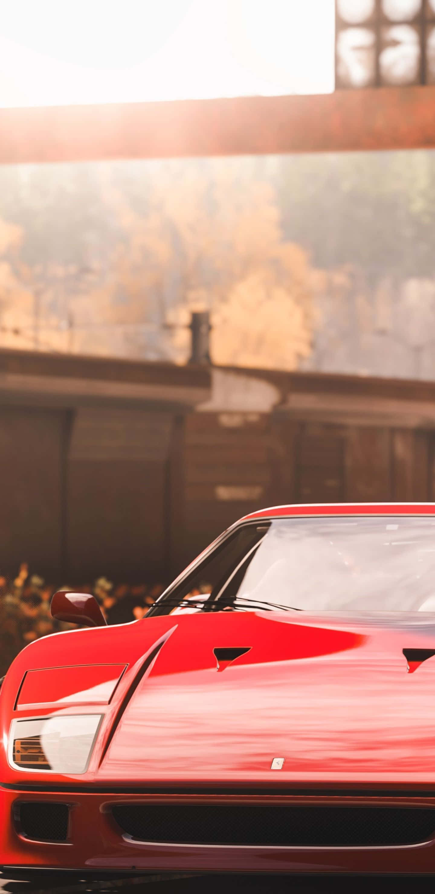 以Pixel 3XL與Forza Horizon 4滿足你對速度的渴望 壁紙 — 一輛光亮的紅色法拉利跑車在溫暖的金色陽光下閃耀，背景是模糊的工業建築和秋天的樹木。