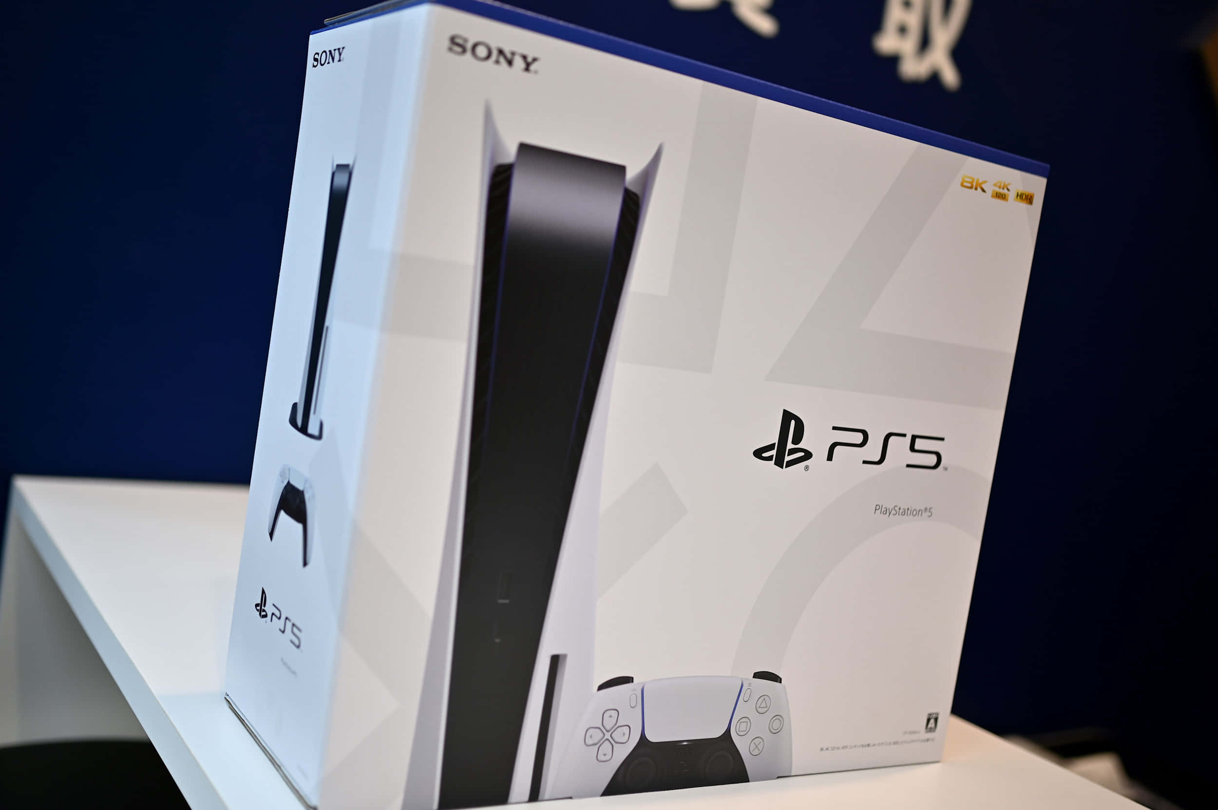 以PlayStation享受全新層次的娛樂遊戲體驗 壁紙 — 一個帶有黑色點綴的白色PlayStation 5主機盒放在白色展示架上，背景為深藍色。
