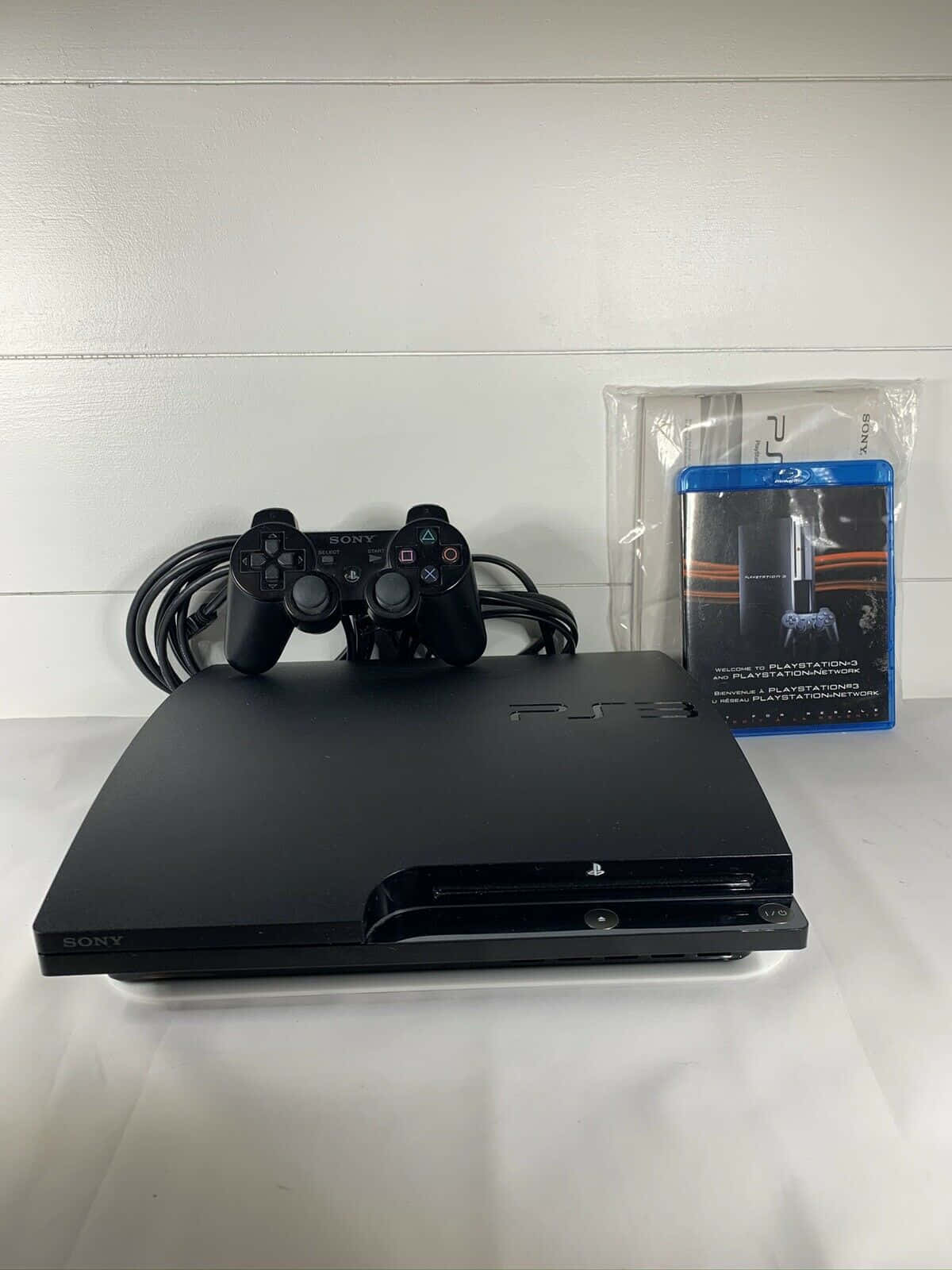 以PlayStation體驗遊戲的下一層次 壁紙 — 一部黑色PlayStation 3主機、手掣及遊戲盒放在白色表面上，背景是白色板牆。