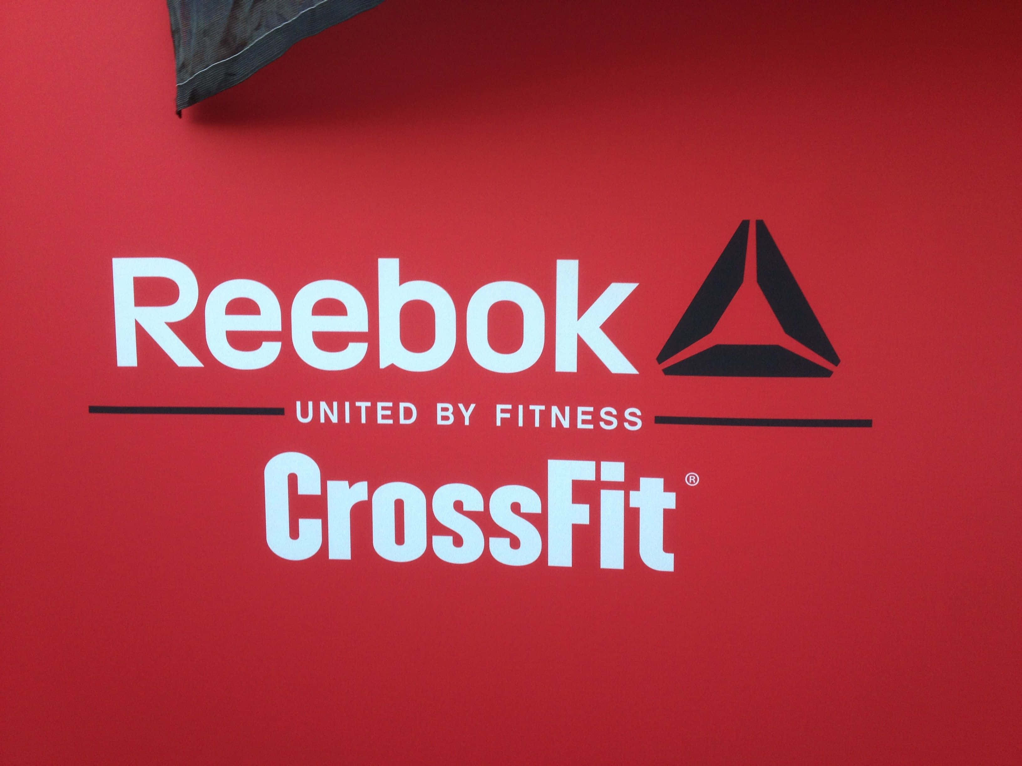 以Reebok突破常規，展現自我 壁紙 — 大膽嘅白色同黑色Reebok CrossFit標誌喺充滿活力嘅紅色牆上，上面有個深色布料角落。