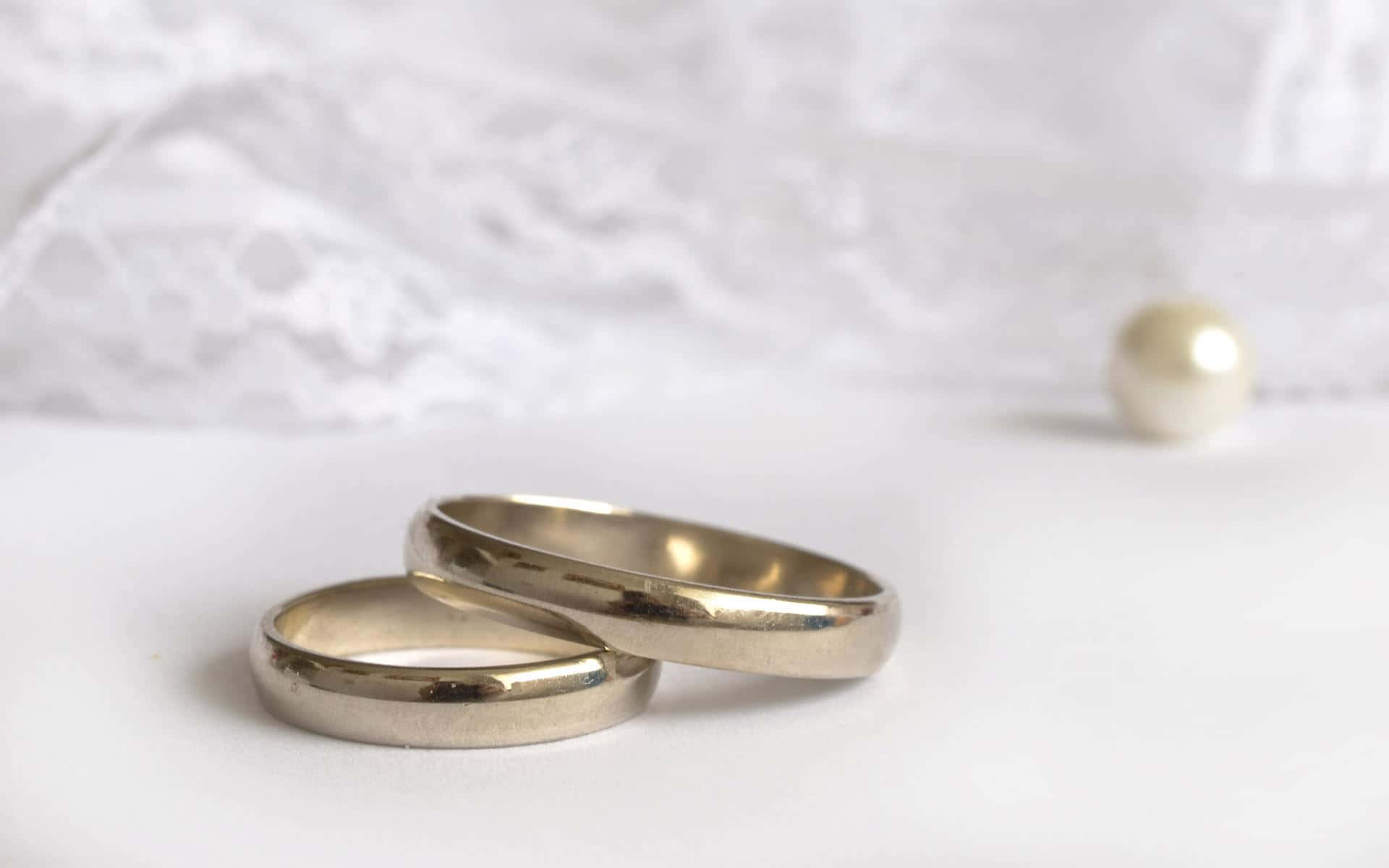以Ring新款智慧珠寶彰顯個性 壁紙 — 兩枚銀色結婚戒指放在白色表面上，旁邊有一顆珍珠，背景是柔軟的蕾絲。