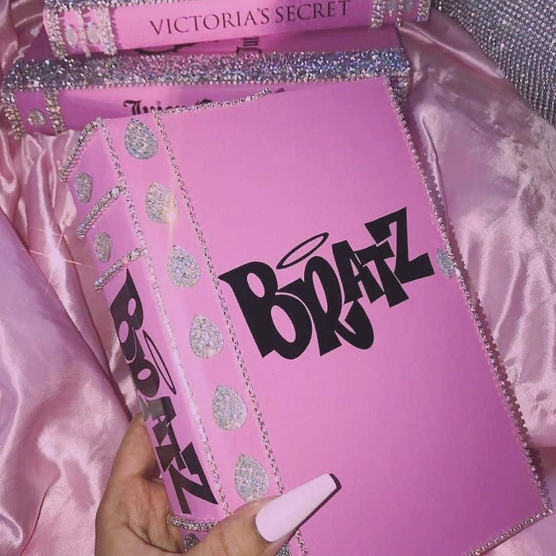 以Y2K美學迎接未來 壁紙 — 一隻手拿著一本閃亮的粉紅色Victoria's Secret書，上面有粗體黑色字母'BOATZ'，背景是緞面粉紅色。