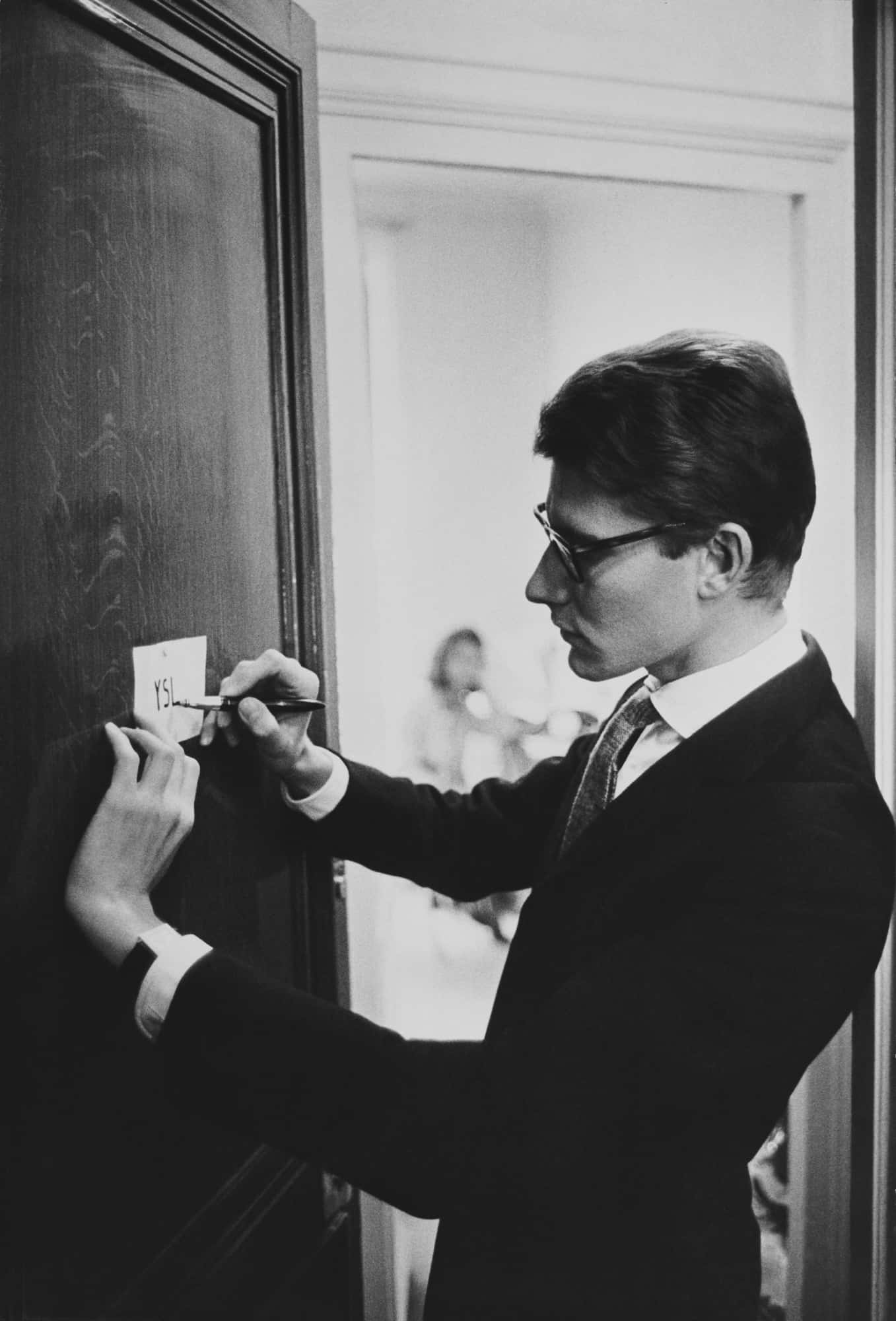 以YSL展現你的大膽個性 壁紙 — 一名身穿西裝、戴眼鏡的男子，在一張黑白照片中，於深色木門上的便利貼寫上「YES」。