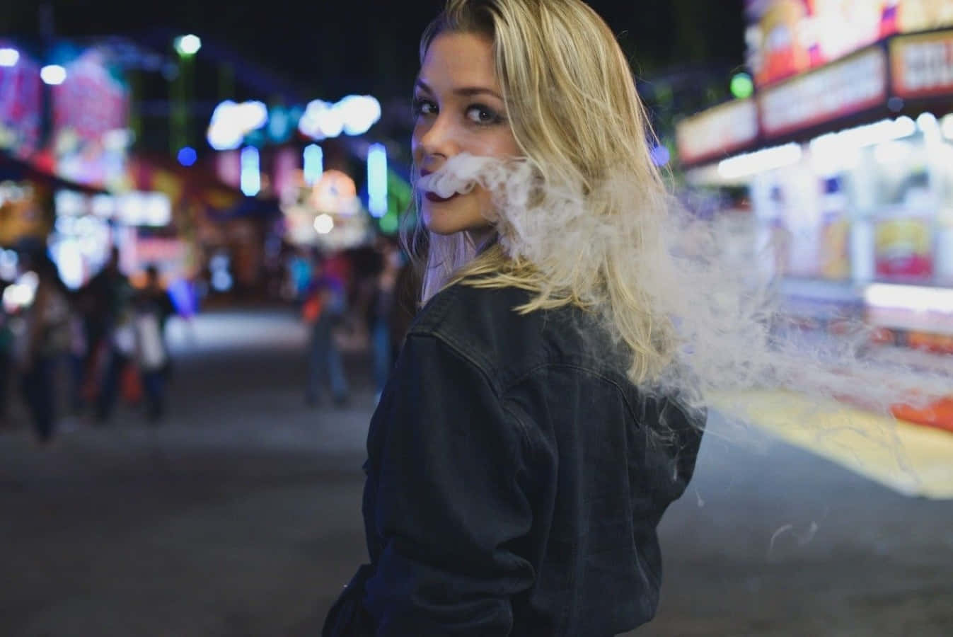 金髮女子在夜晚霓虹燈閃爍的都市街道上呼出煙霧。