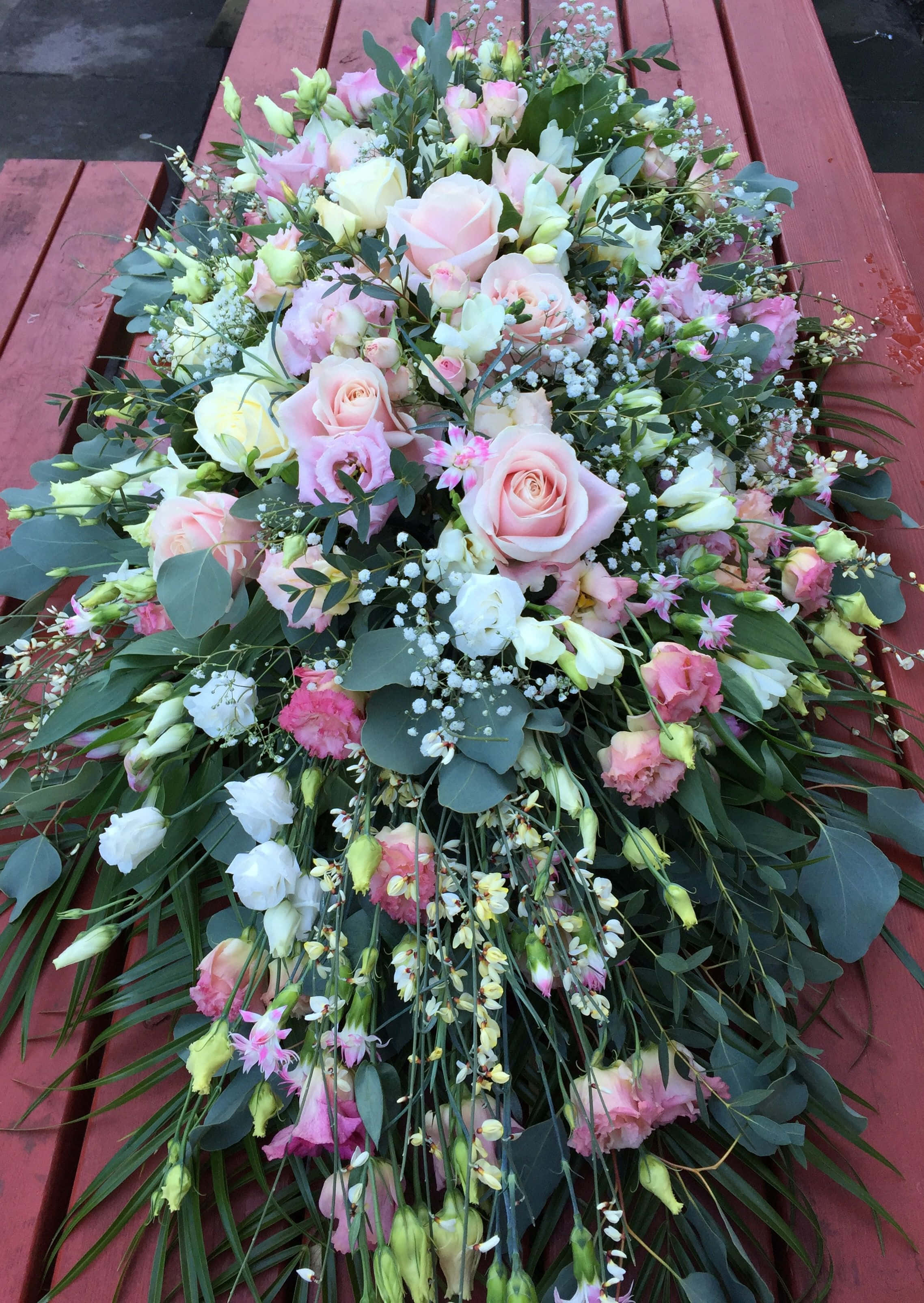 以優雅喪禮花束表達哀悼 壁紙 — 粉紅玫瑰、滿天星和綠植組成的茂密瀑布，優雅地擺放在一條風化的紅色木長椅上。