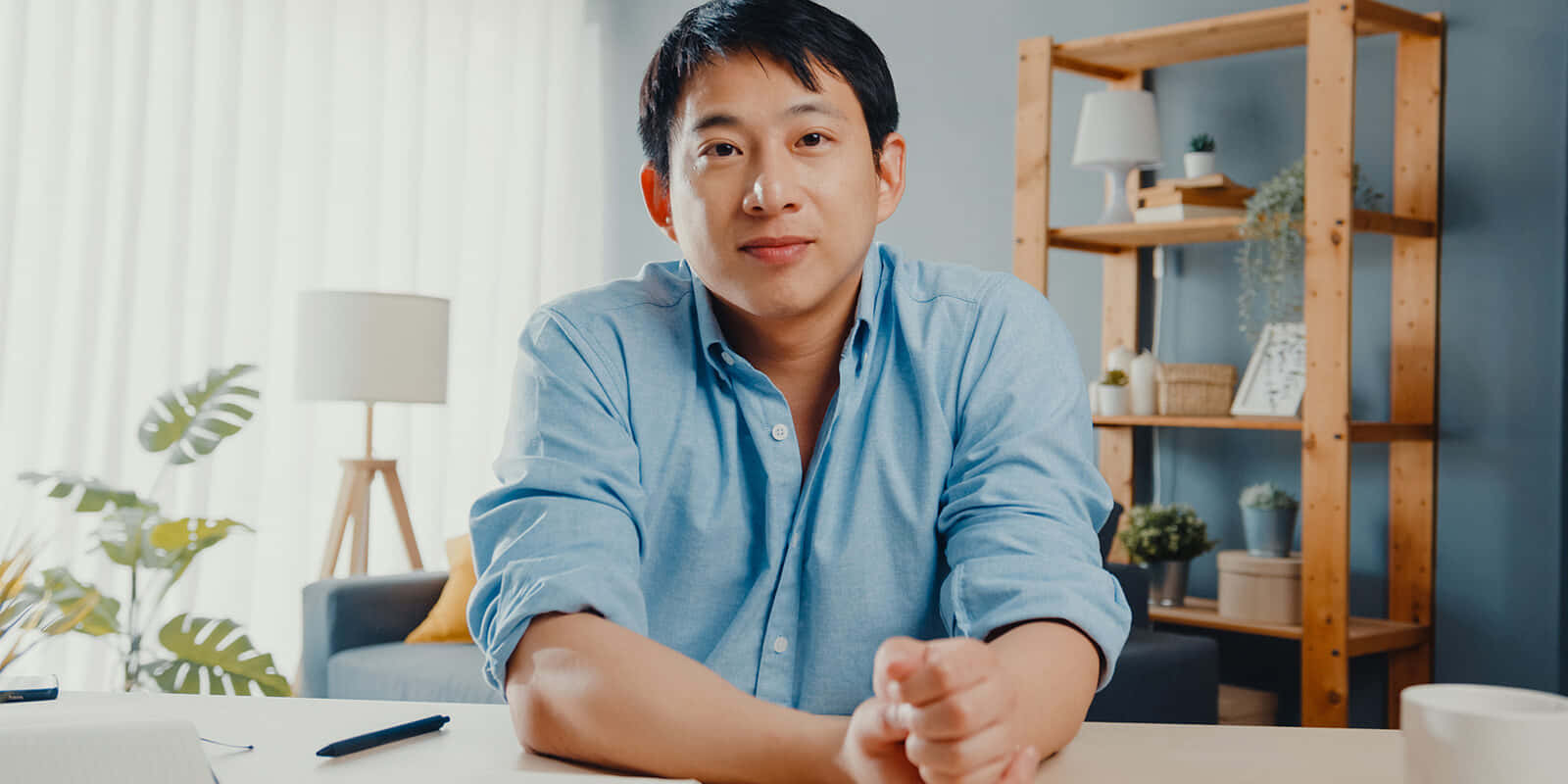 以優雅桌面壁紙點亮您的空間 壁紙 — 一位穿著藍色襯衫的冷靜男子坐在一個有植物和架子的舒適家庭辦公室的木桌旁。