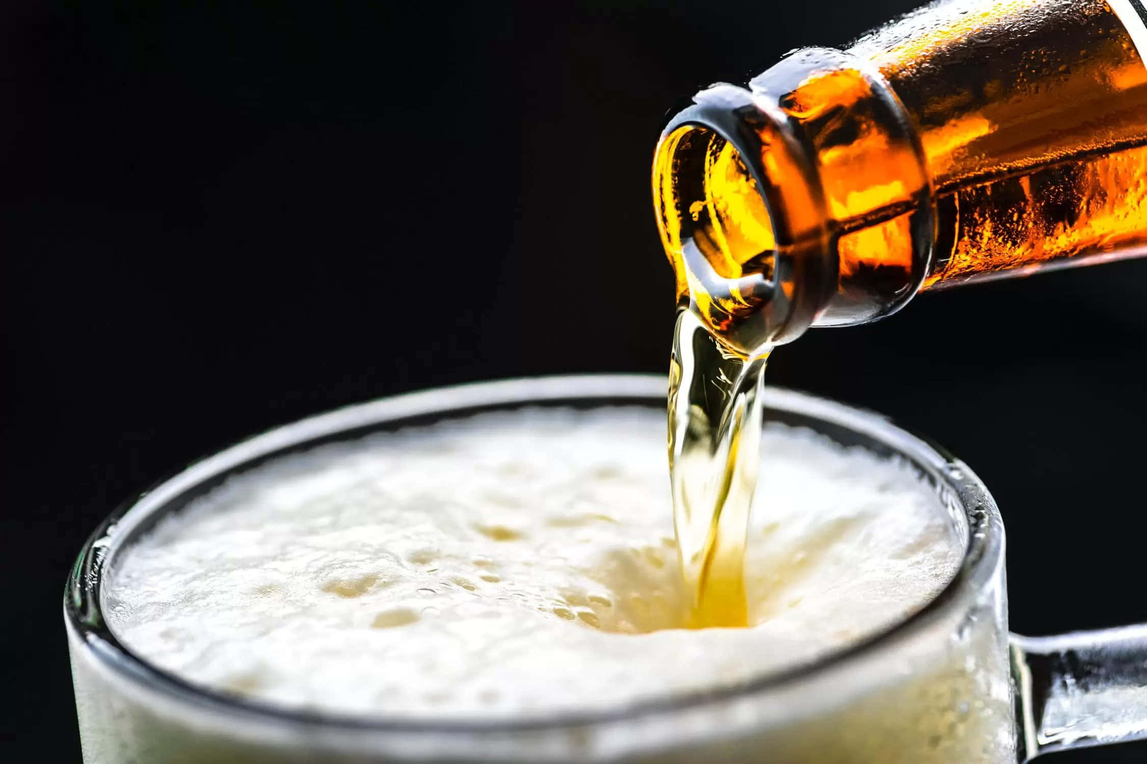 以冰啤酒慶祝 壁紙 — 金黃色啤酒從冰凍的琥珀色瓶子倒入玻璃杯中，在深色背景下形成泡沫豐富的白色泡沫。