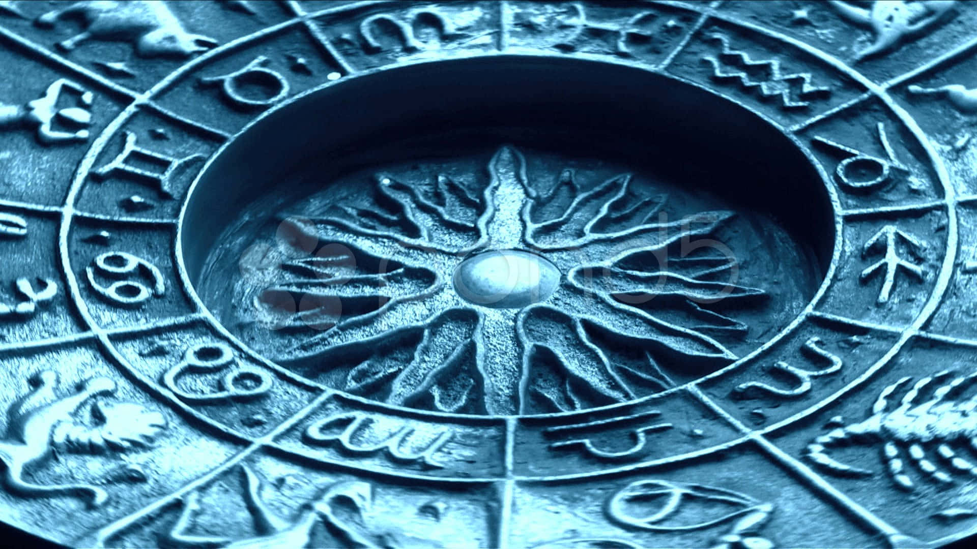 以占星學探索宇宙未知 壁紙 — 古代占星黃道輪的微距特寫，帶有雕刻符號和中央太陽爆發圖案，在冷藍色燈光下。