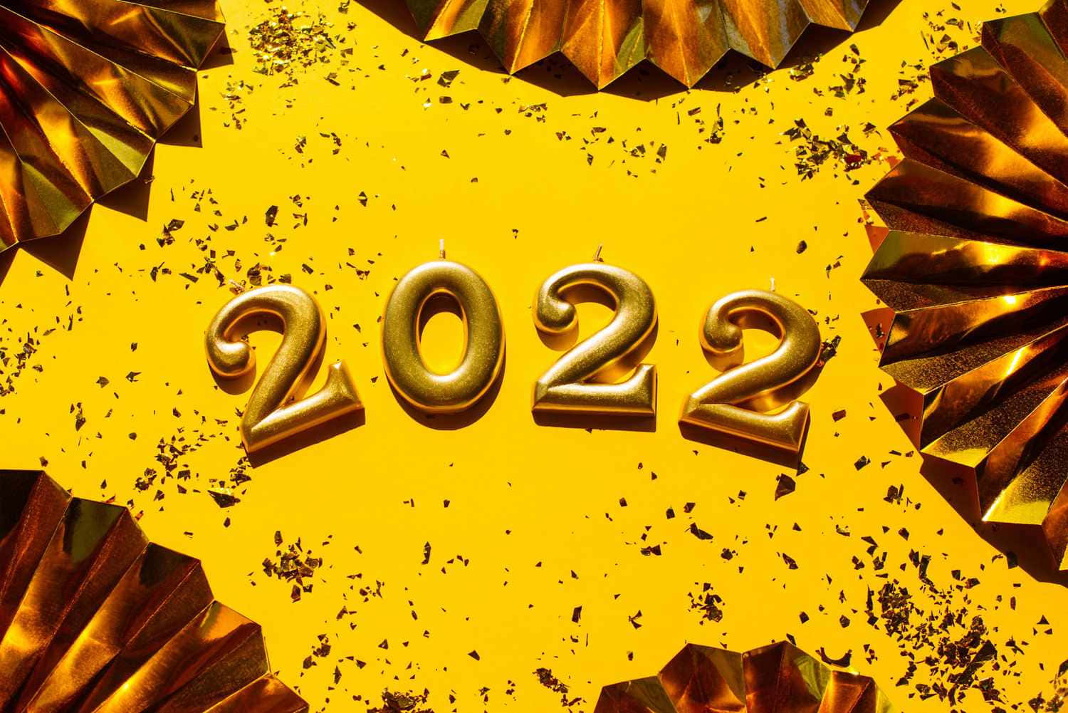 以喜悅與煙火迎接新年！ 壁紙 — 金色2022蠟燭放在明亮的黃色背景上，周圍是金屬扇裝飾和五彩紙屑。
