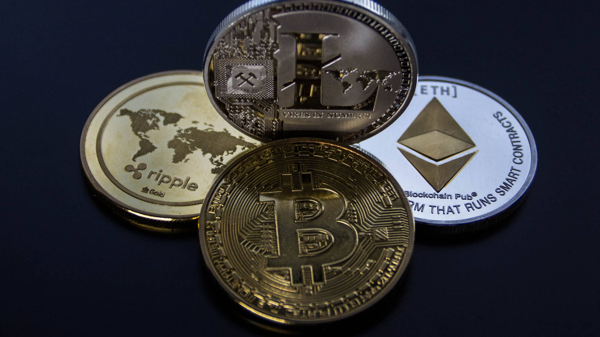以太幣加密貨幣排名 壁紙 — 四個實體加密貨幣硬幣 Bitcoin Ethereum Ripple 和 Litecoin 排列在帶有金屬光澤和雕刻標誌的深色表面上。