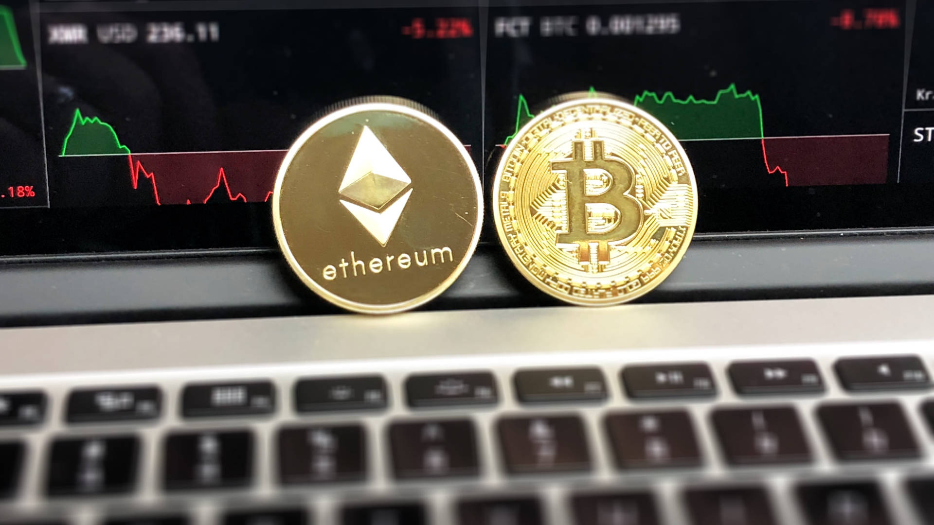 以太幣與比特幣在筆記本電腦上 壁紙 — 兩枚金色加密貨幣以太坊和比特幣放在筆記型電腦鍵盤上，背後有股市圖表發光。