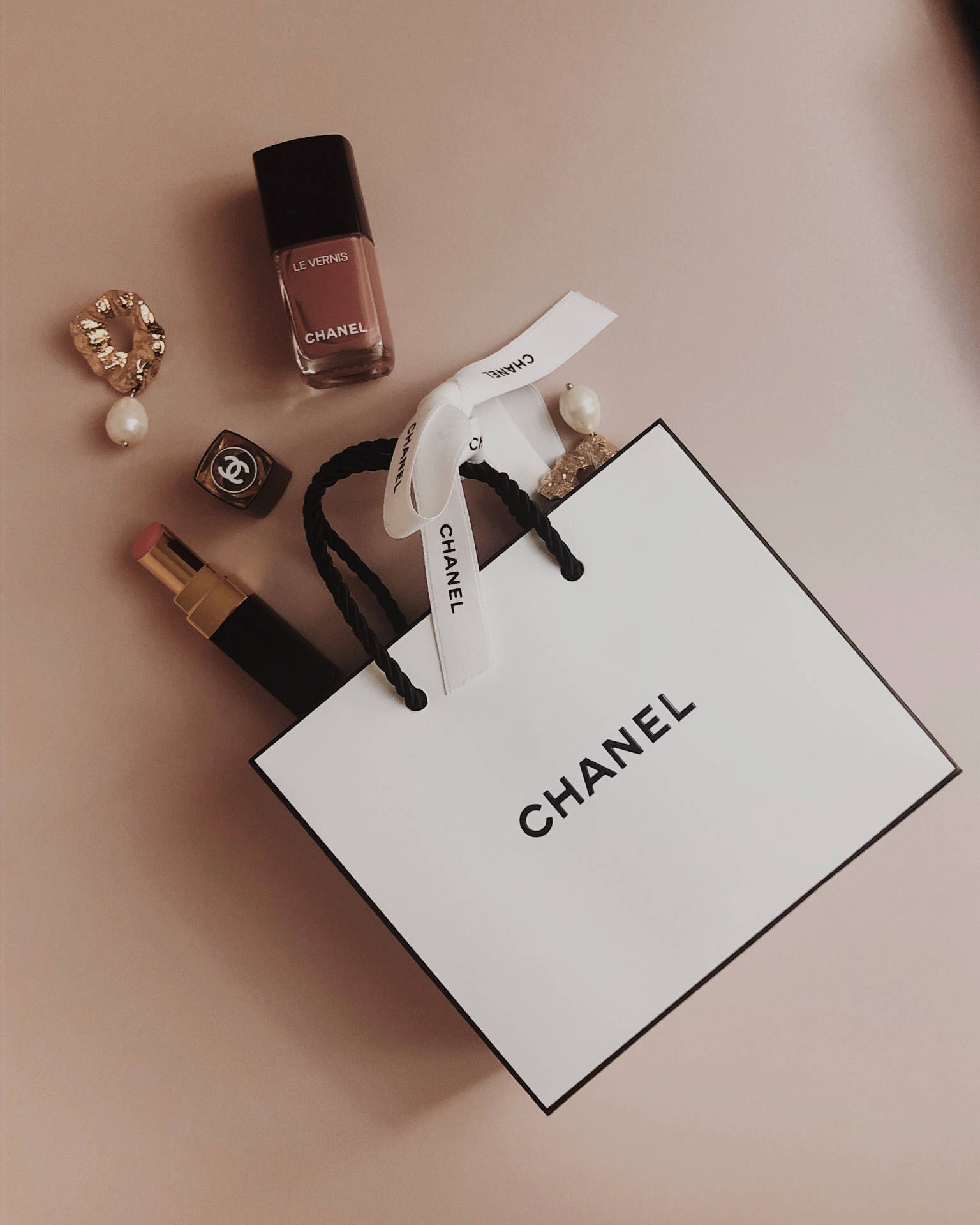 Chanel 購物袋搭配指甲油、唇膏和珠寶，置於粉紅色表面上。